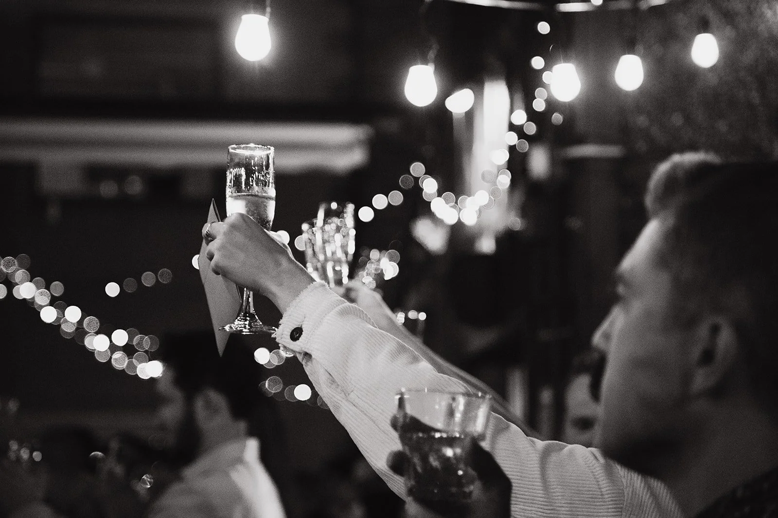 A person raising a glass of champagne during a celebration in a dimly lit environment with string lights.