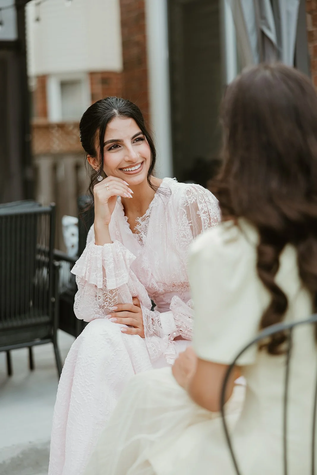Two women sitting outdoors, engaged in conversation, with one wearing a white lace dress and smiling, the other with long brown hair in a cream dress at an engagement party in Mississauga, Ontario.