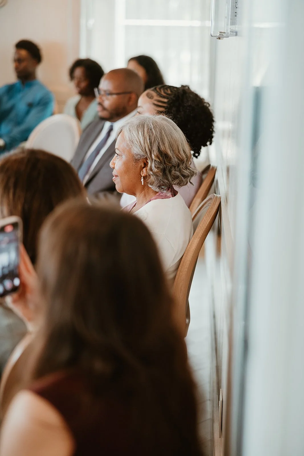 People sitting in a row attending an indoor event, some using their phones, in a well-lit room with large windows and sheer curtains at Katb al Kitab at the Pearle Hotel in Burlington, Ontario.