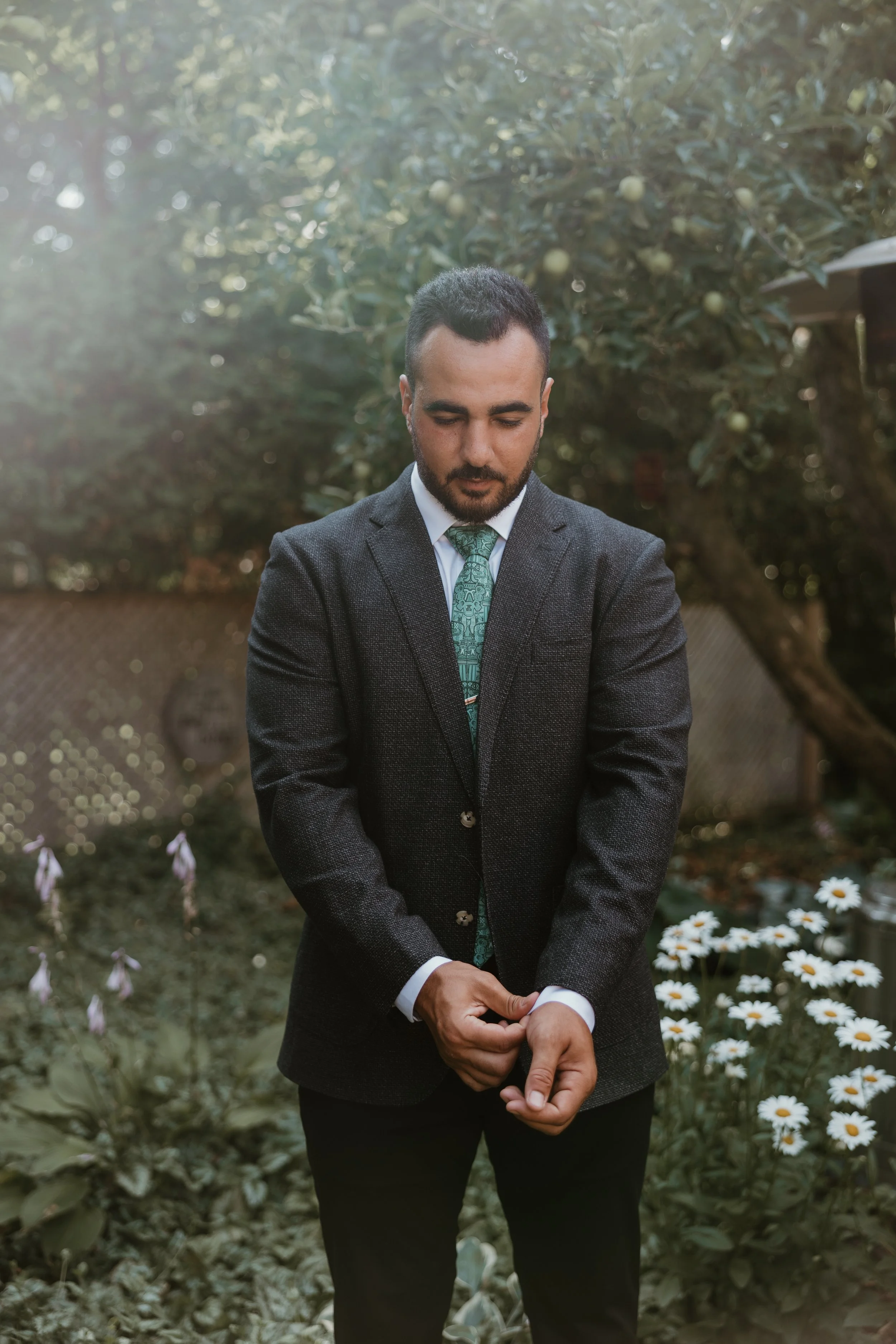 A man wearing a dark suit and a patterned tie standing outdoors among flowers and greenery, with a contemplative expression and his hands adjusting his sleeve, at a Katb al Kitab in Guelph, Ontario.