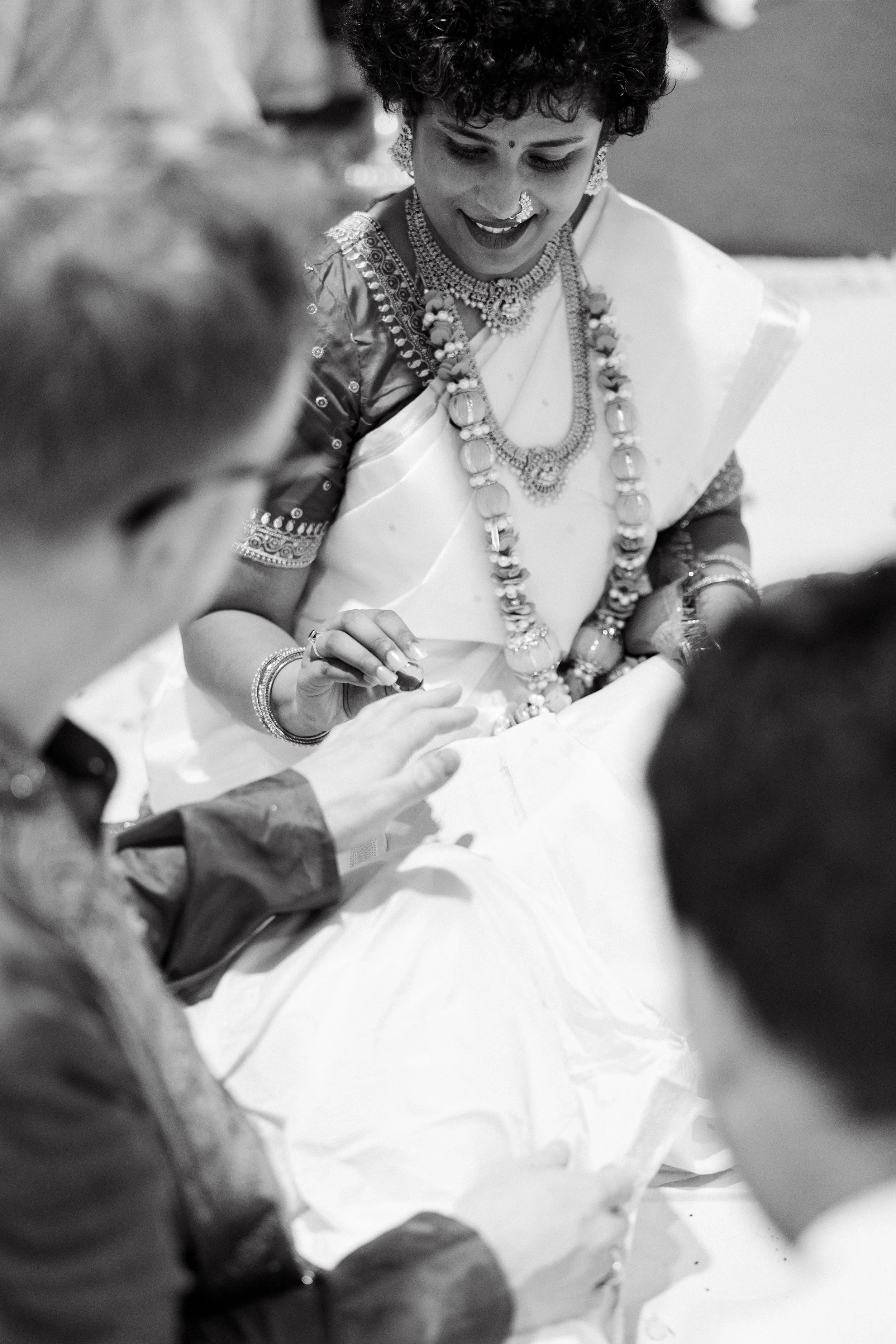 A woman wearing traditional Indian jewelry and a saree, smiling and looking at her hand, with blurred people around her, in a black and white photo, at a traditional wedding at a temple in North York, Ontario.