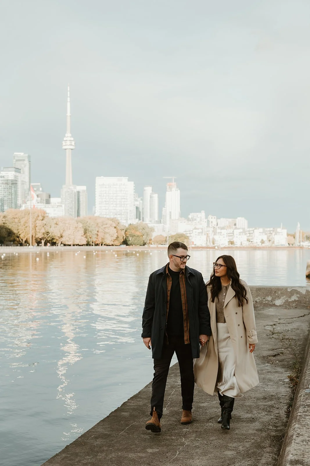 A couple walking along a waterfront promenade in Toronto, with the Toronto skyline and CN Tower in the background, during daytime.