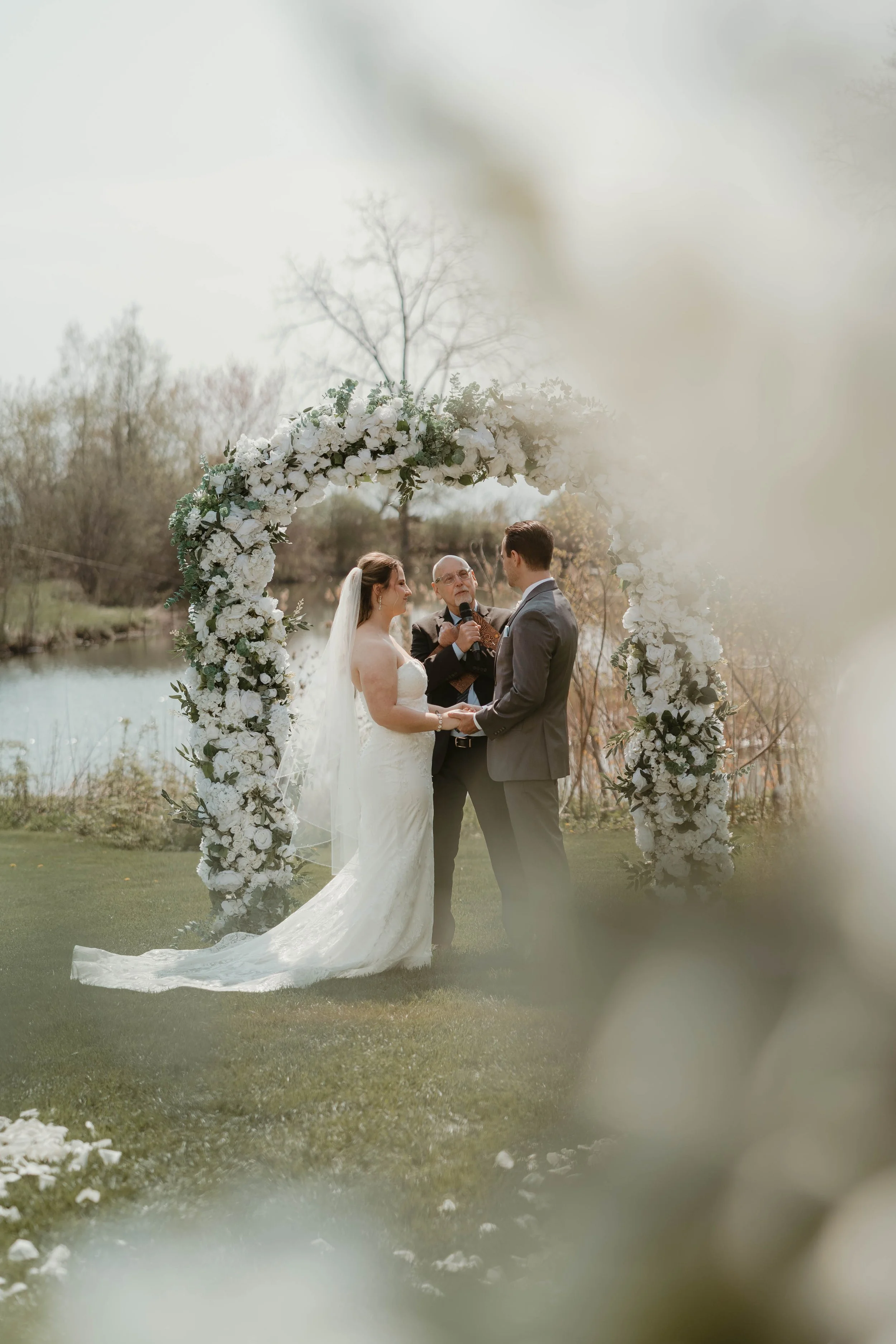 A bride and groom exchange vows under a floral arch during an outdoor wedding ceremony by a lake, with officiant present on a cloudy day at Harding Estate in Oakville, Ontario.