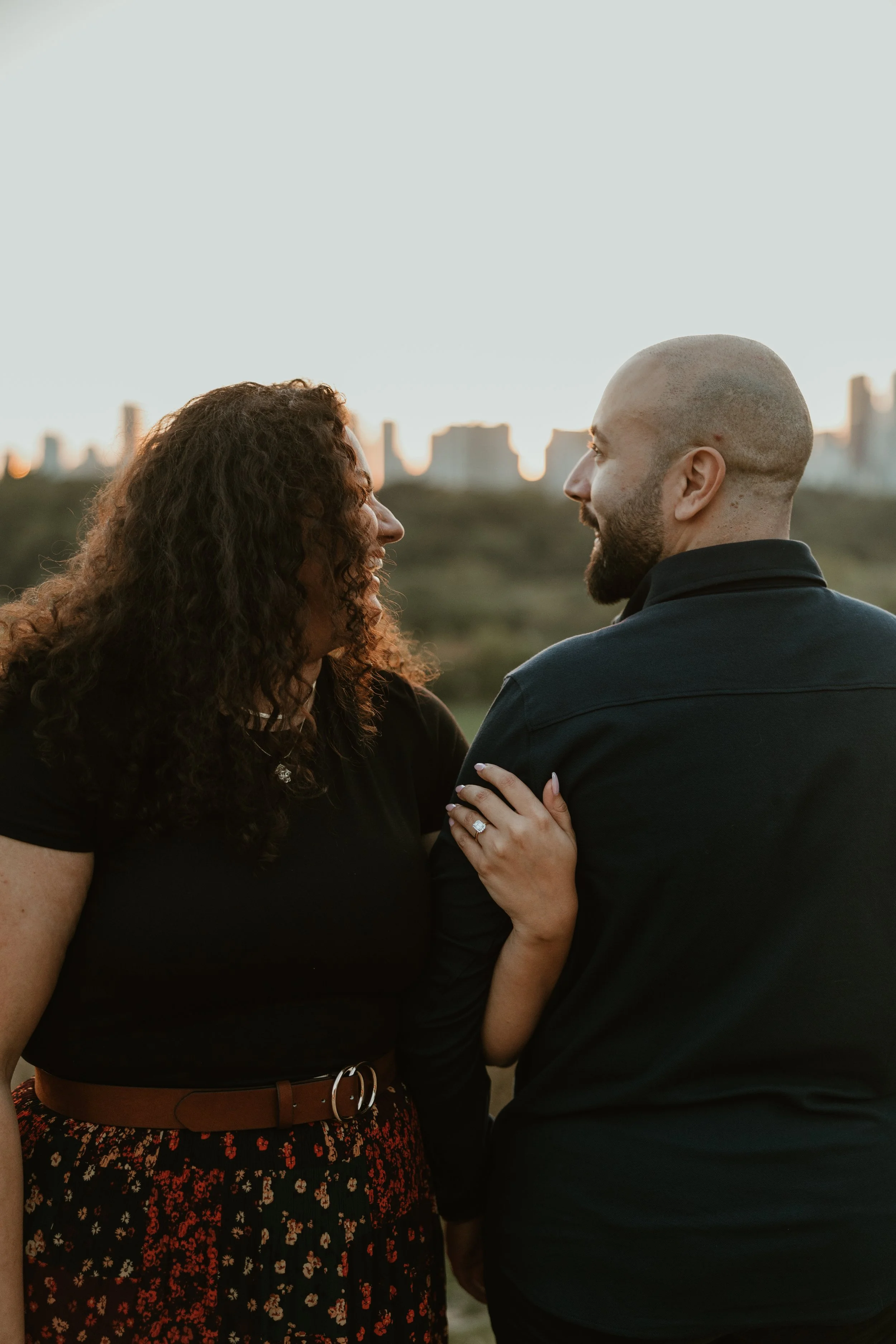 A couple gazing at each other outdoors during sunset, with city skyline in the background after their proposal in Riverside Park in Toronto, Ontario.