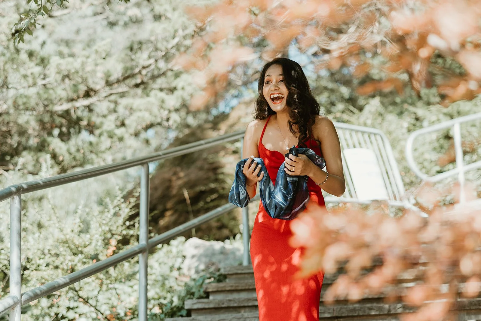 A woman in a red dress smiling and walking down outdoor stairs surrounded by greenery and autumn leaves for her proposal in Edwards Gardens in Toronto, Ontario.