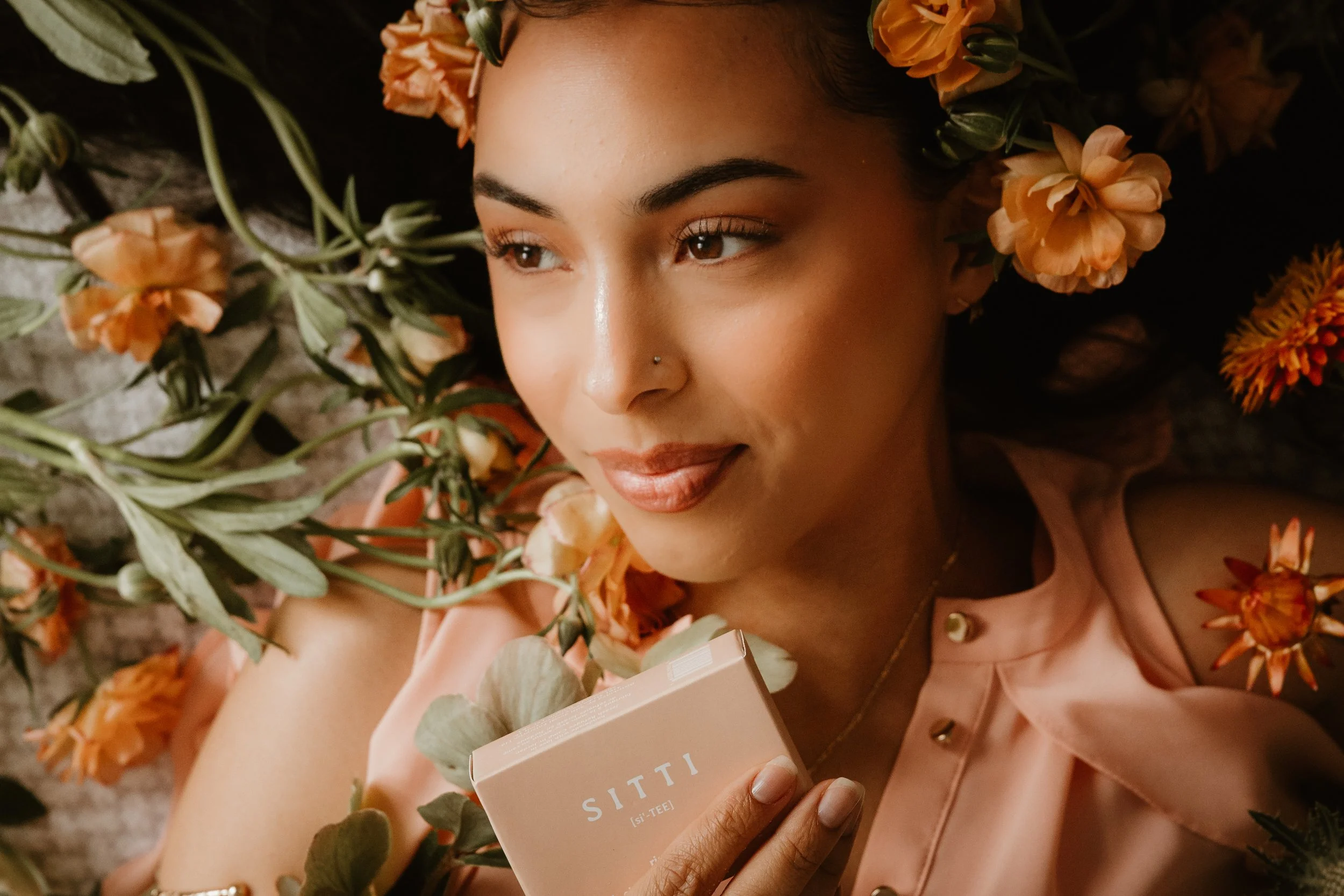 A woman lying down surrounded by orange flowers, holding a pink box, with a content expression.
