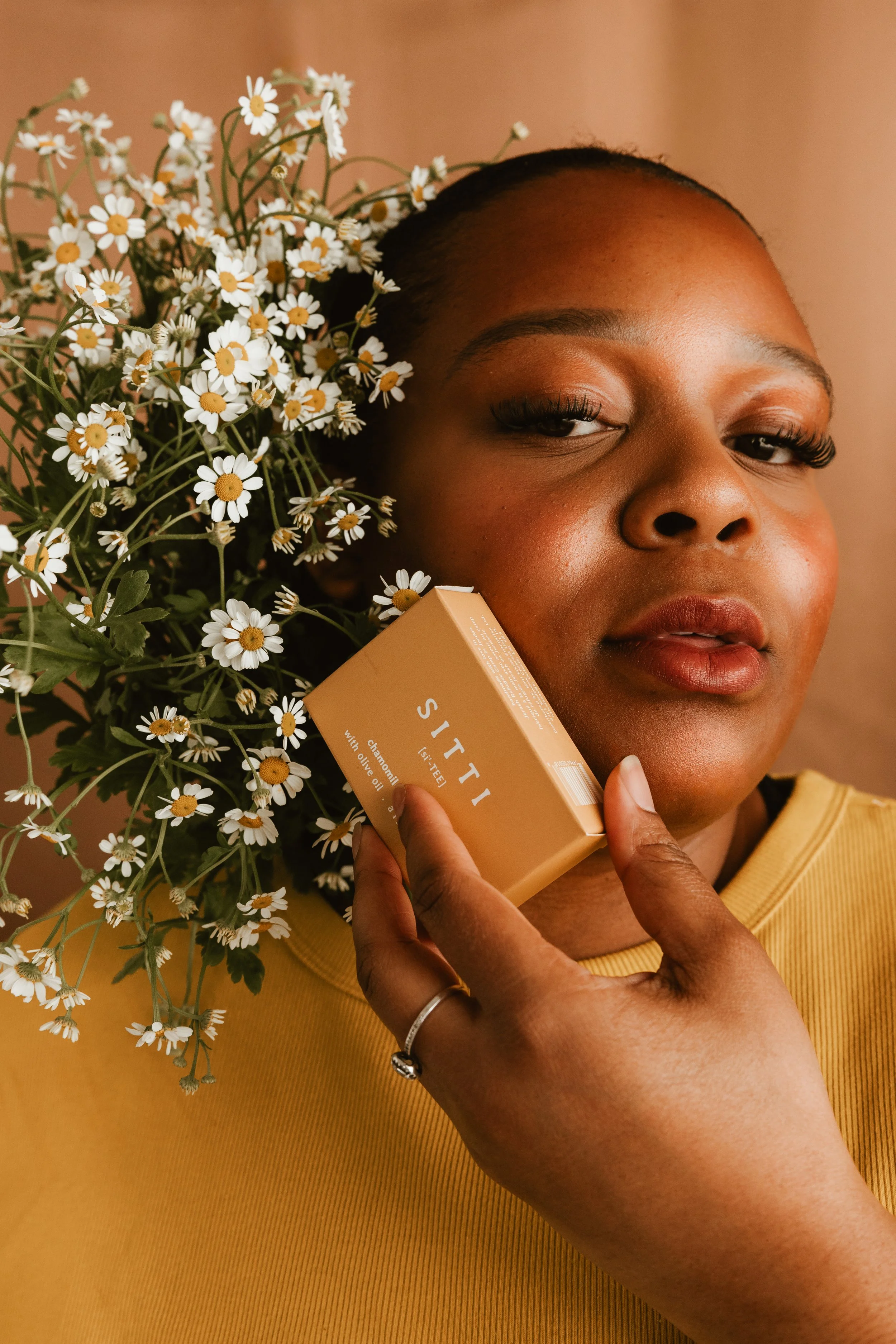 A woman with dark skin and natural hair poses with a bouquet of daisies and holds a beige box labeled 'SITTI' close to her face, wearing a yellow top.