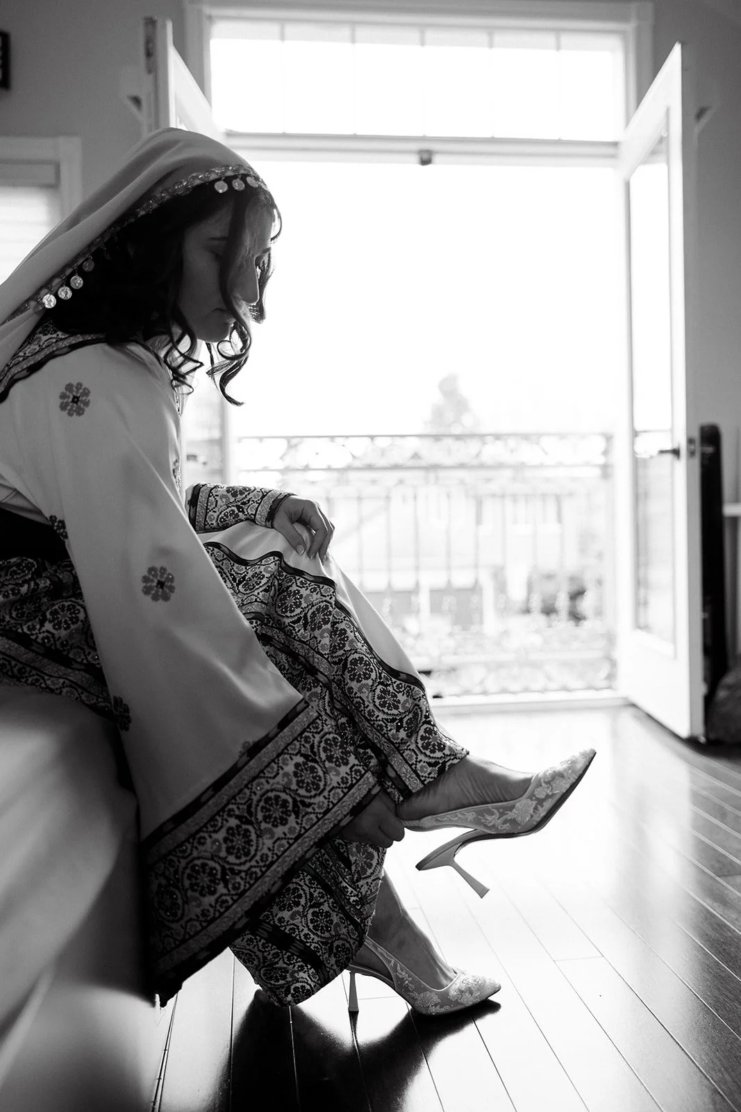 A woman in a traditional embroidered dress sitting on a bed, putting on elegant high-heeled shoes, with an open door and balcony in the background, getting ready for her wedding in Oakville, Ontario.