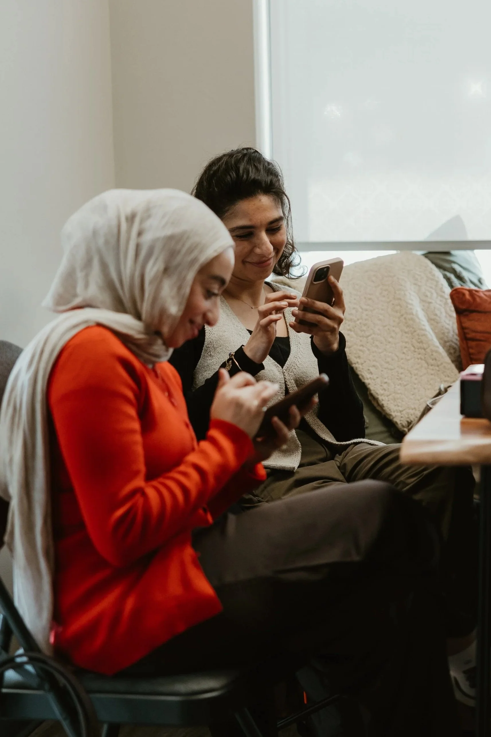 Two women sitting on a couch, smiling and looking at their phones. One woman is wearing a hijab and orange top, and the other woman has short curly hair and is wearing a beige and black jacket at Sitti Soap in Mississauga, Ontario.