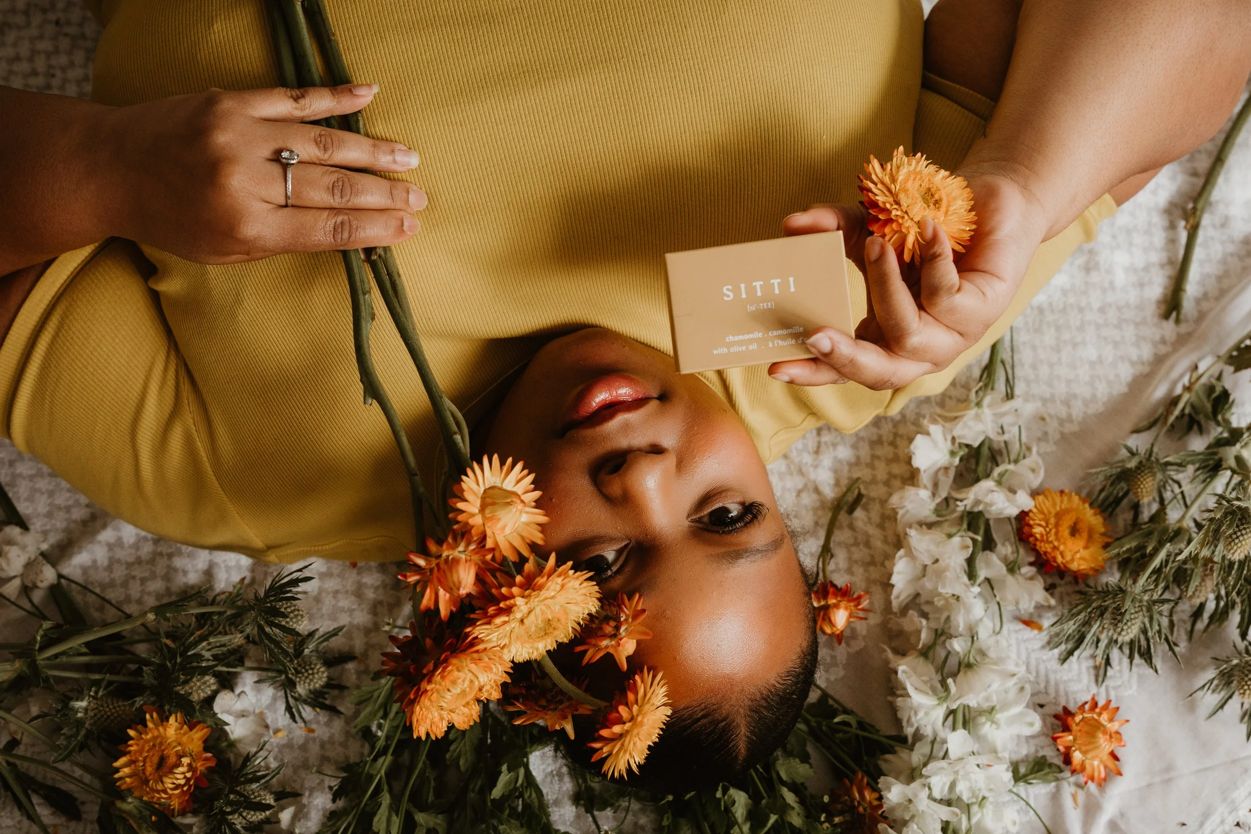 A woman lying on the floor surrounded by flowers, holding a beige box labeled 'SITTI' and a flower, with her eyes closed and wearing a yellow outfit.