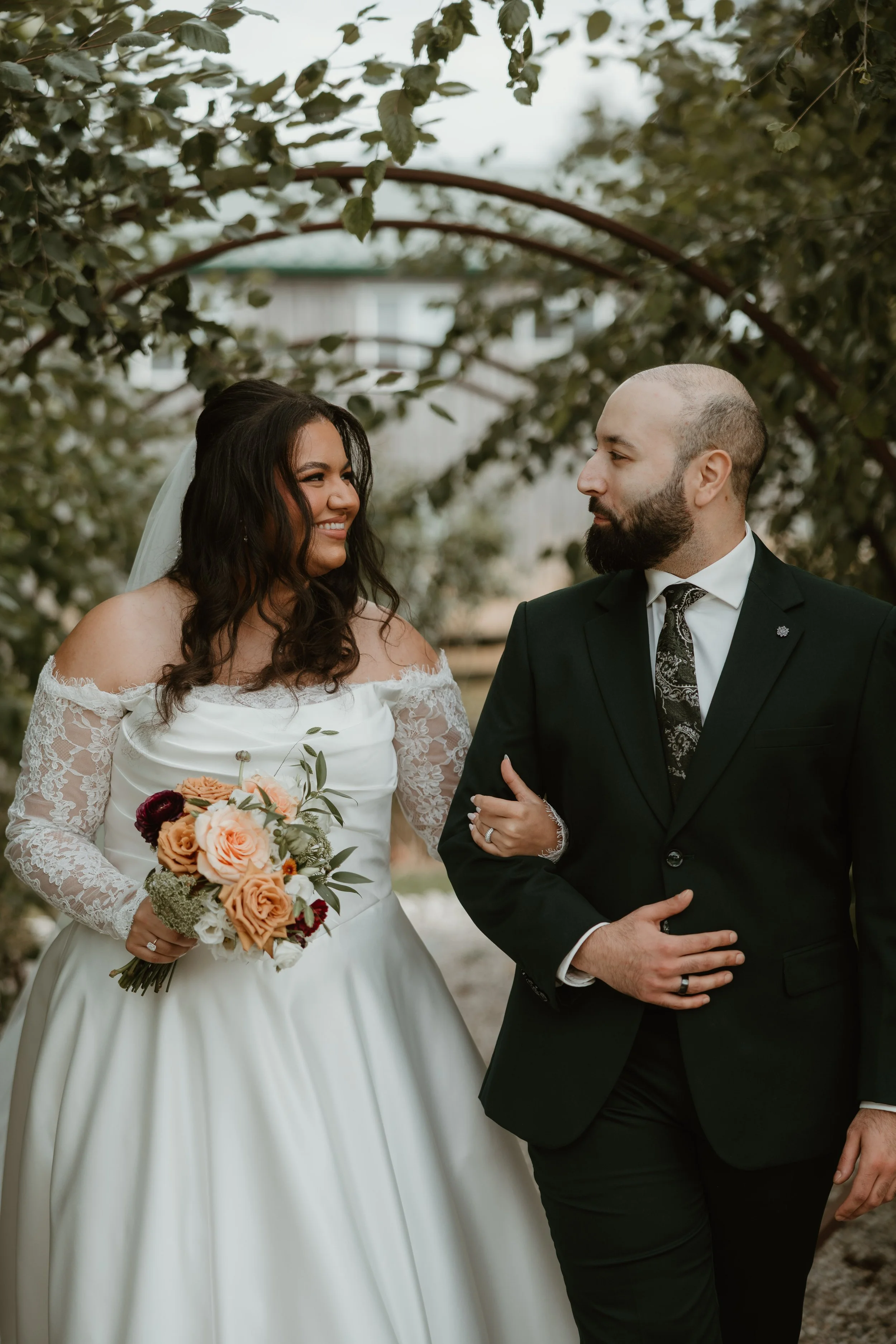 A bride and groom standing outside in front of trees, looking at each other and smiling. The bride wears a white wedding dress with lace sleeves and holds a bouquet of orange, white, and red flowers. The groom wears a black suit with a patterned tie and has a beard.