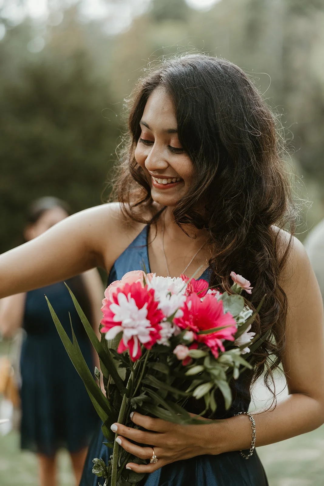 A woman with long, curly dark hair, smiling and looking down, holding a bouquet of pink and white flowers, wearing a sleeveless dark blue dress and a bracelet, outdoors with a blurred background in Edwards Gardens in Toronto, Ontario.