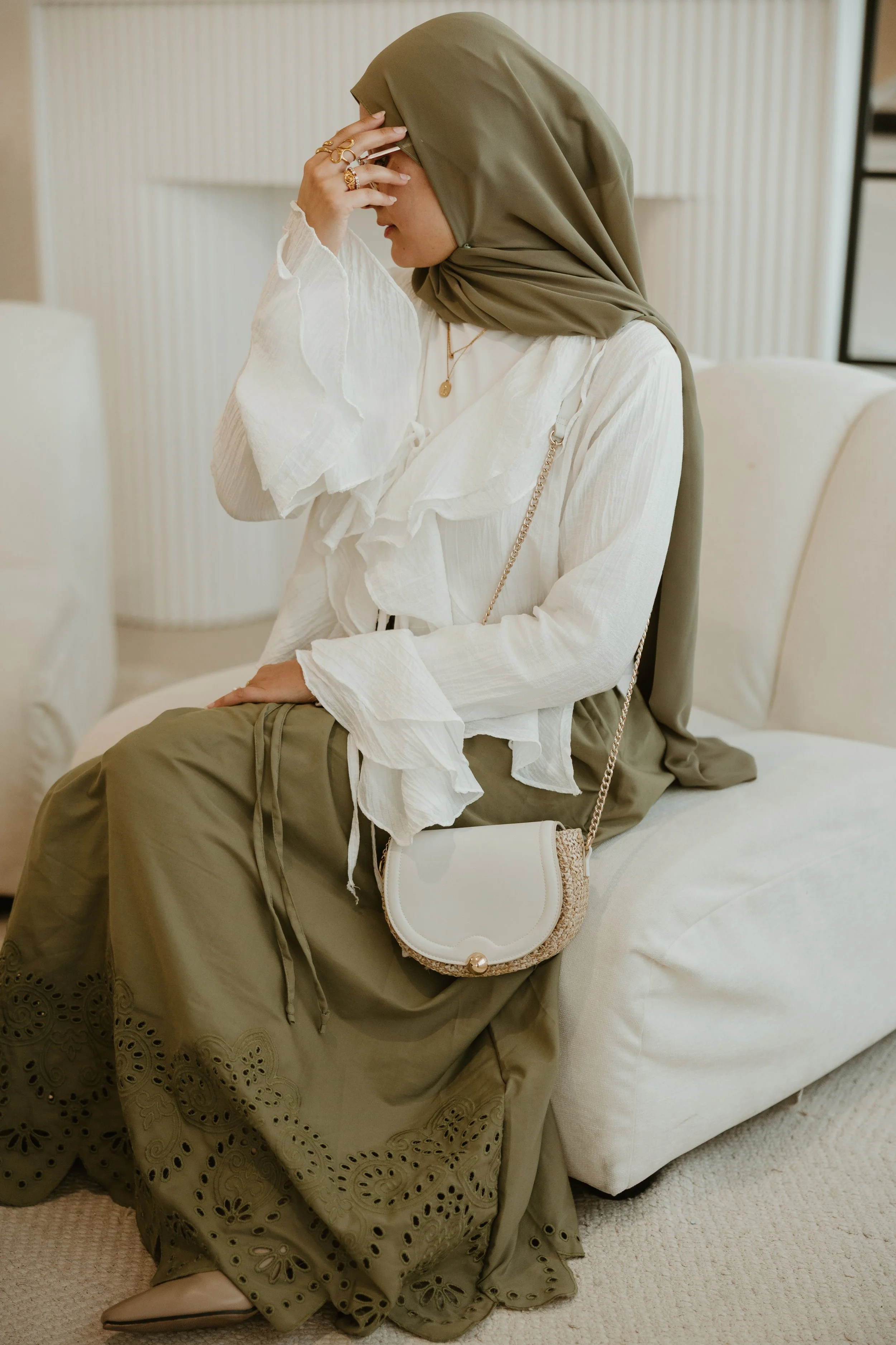 A woman wearing a green hijab and a white ruffle blouse, sitting on a white cushion, holding her head in her hand with rings on her fingers, a necklace, a white purse with a chain strap, and a long olive green skirt with cut-out floral designs.  background shows a beige wall and a black metal shelf.
