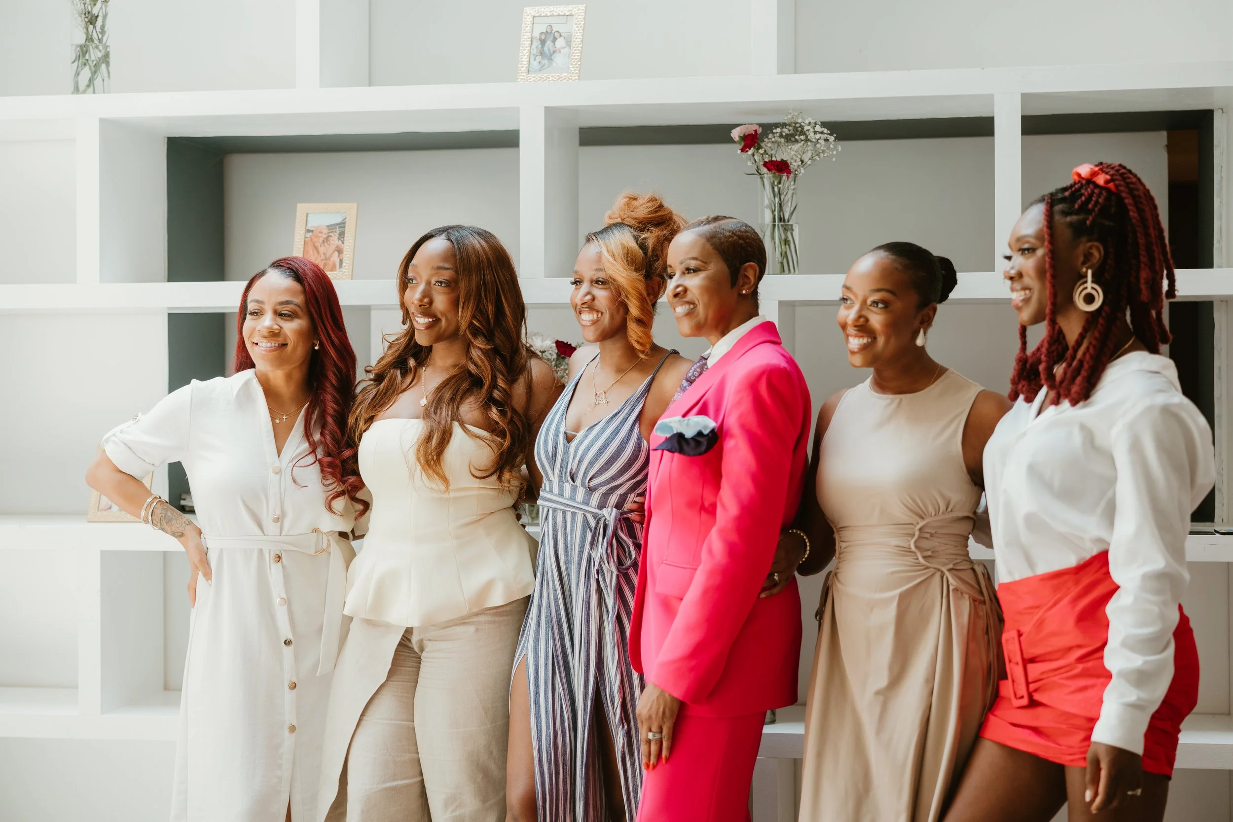 Six women standing together in a row, smiling, in front of white shelving with framed photos and flowers.