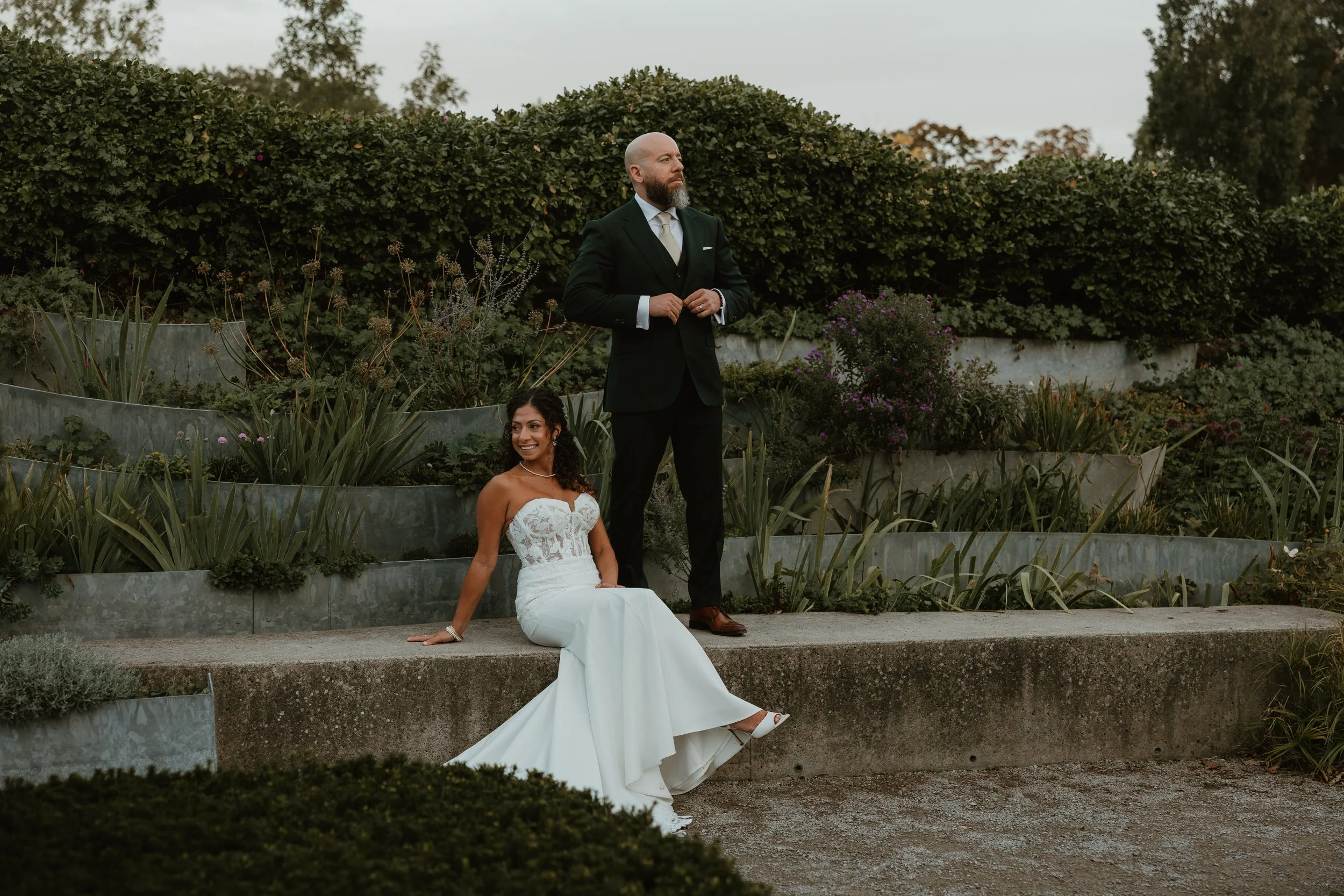 A bride in a white wedding gown sitting on a concrete ledge outdoors, smiling, with a columnist man standing behind her with his hands clasped, both in front of dense greenery and flowers.