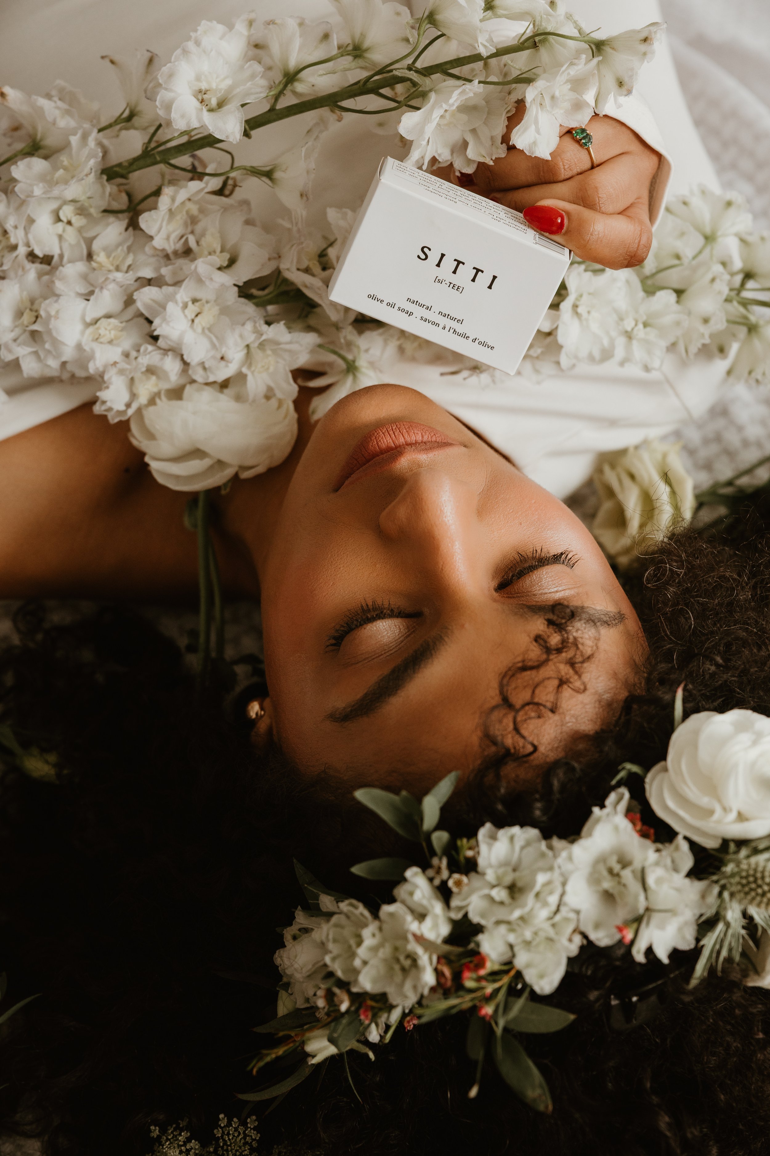 A woman with curly hair and a ring is lying down amidst white flowers, holding a box of SITI olive oil soap, with her eyes closed and a peaceful expression.