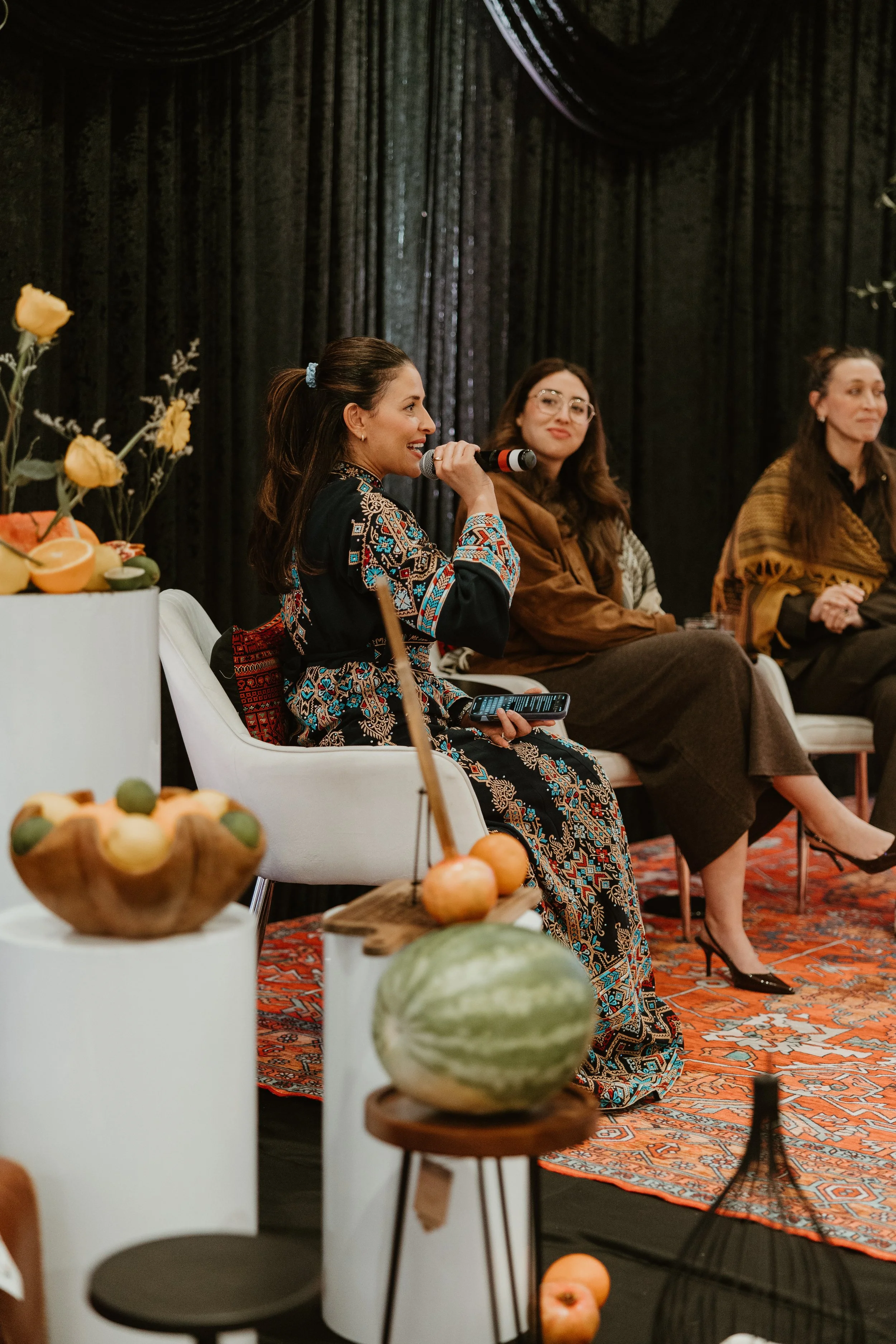 Women sitting on chairs in front of black curtains, with a woman speaking into a microphone, and food and fruit displays in the foreground, at Oakville Legacy Banquet Hall.