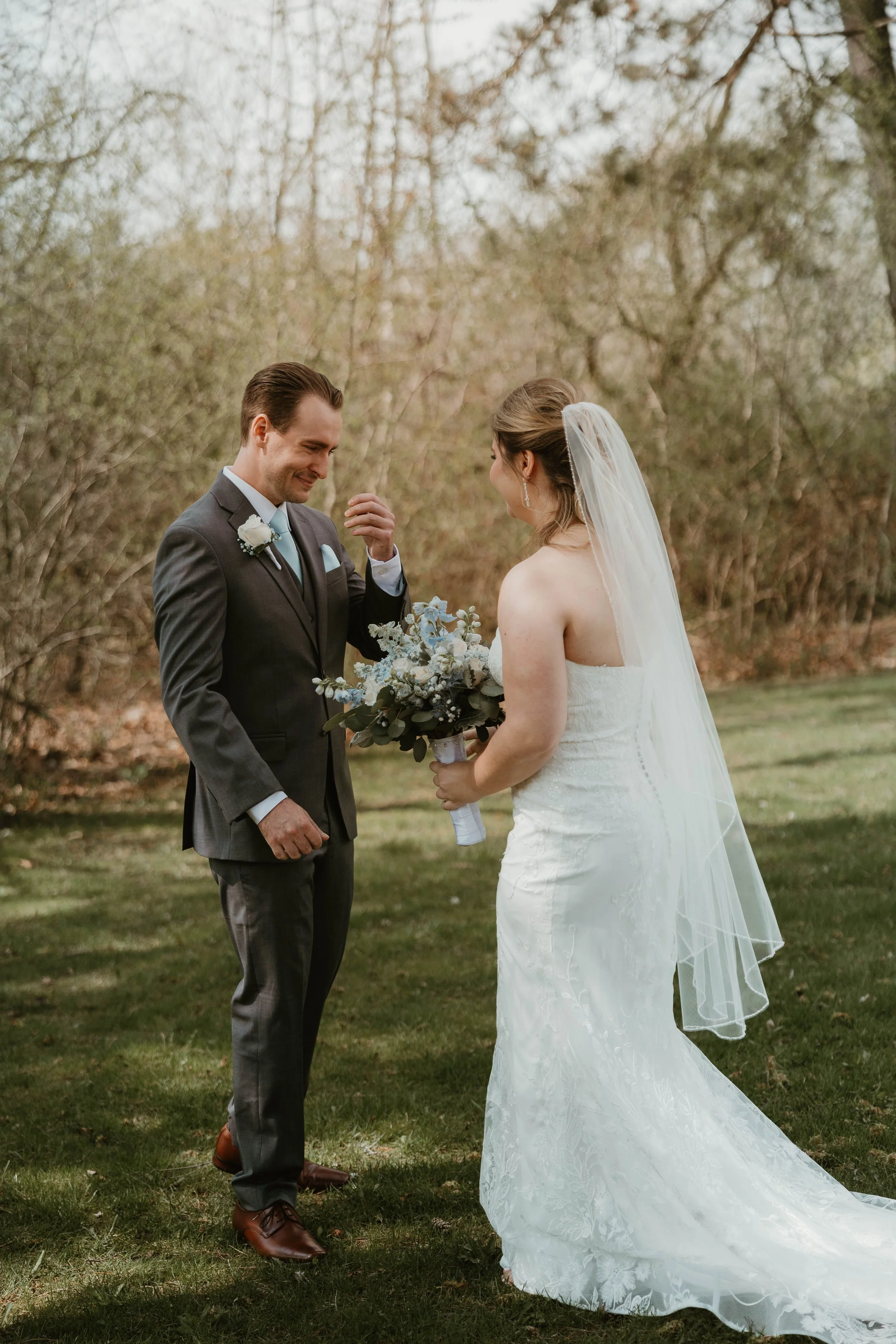 A groom in a grey suit with a white boutonniere and brown shoes looks at his bride, who is wearing a strapless wedding gown with a long veil, on a grassy outdoor area with trees in the background, at Harding Estate in Oakville Ontario