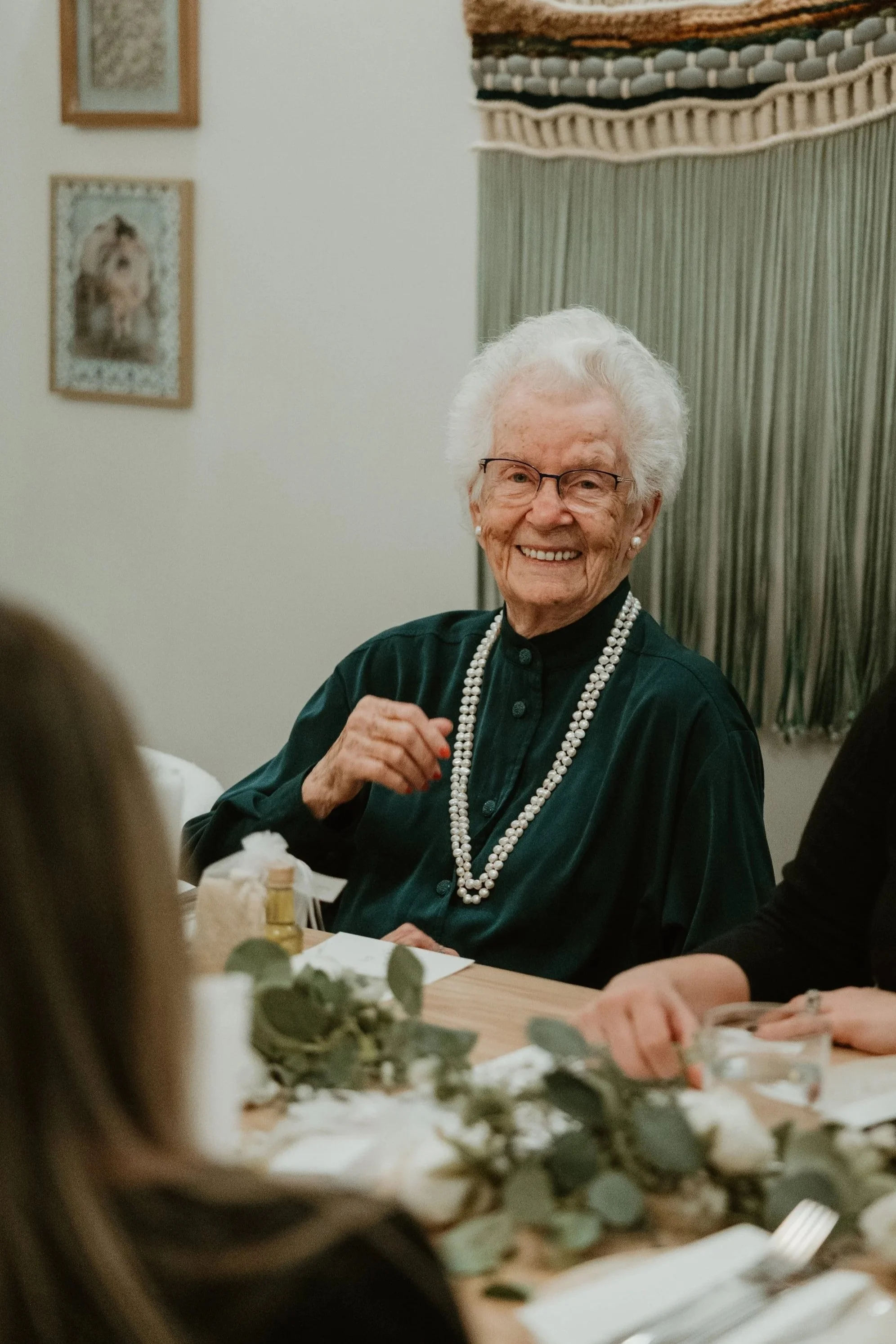 An elderly woman with white hair, glasses, and pearl earrings smiling while sitting at a table during a gathering or celebration, at a wedding reception at Tabule in Oakville, Ontario.