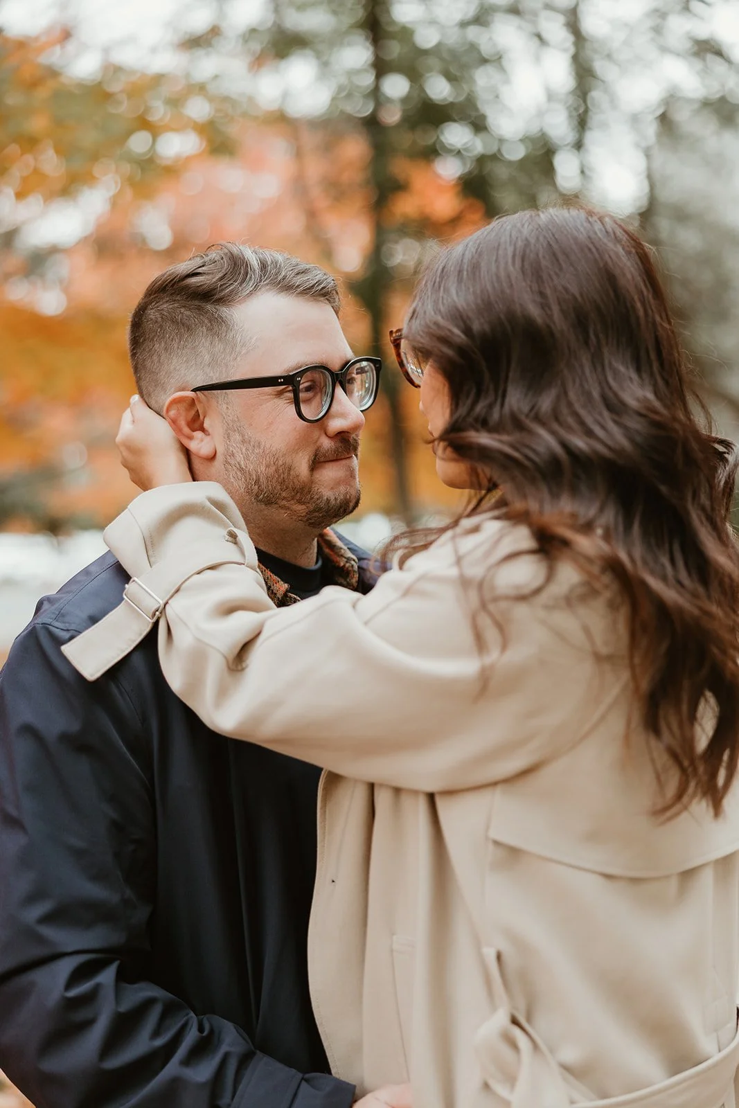 A man and woman face each other closely outdoors during autumn, with blurred orange and yellow fall leaves in the background.