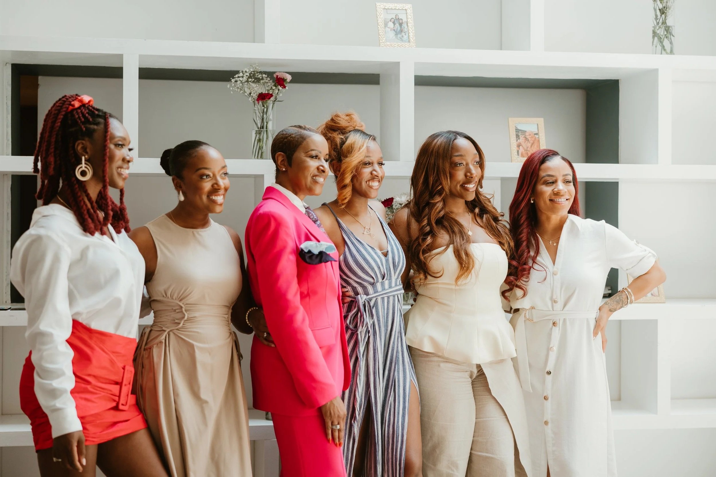 Six women standing together indoors, smiling for a group photo in front of white shelves with decorative items, including framed pictures and vases with flowers.