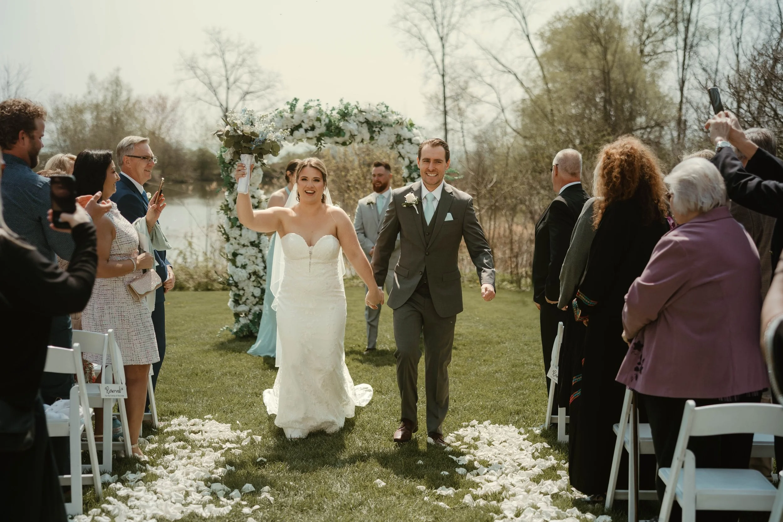 A bride and groom are walking down the outdoor wedding aisle, surrounded by seated guests, with a floral arch in the background on a sunny day at Harding Estate in Oakville, Ontario