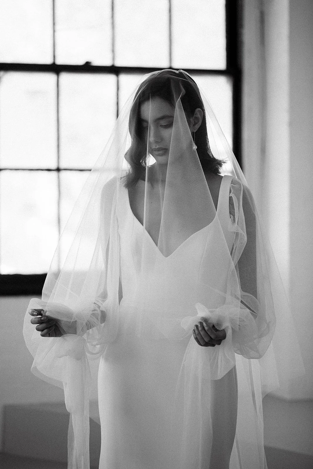 Black and white photo of a woman wearing a wedding dress and veil, standing near a large window, looking down with a serene expression at Park Hill Studios in Cambridge, Ontario.