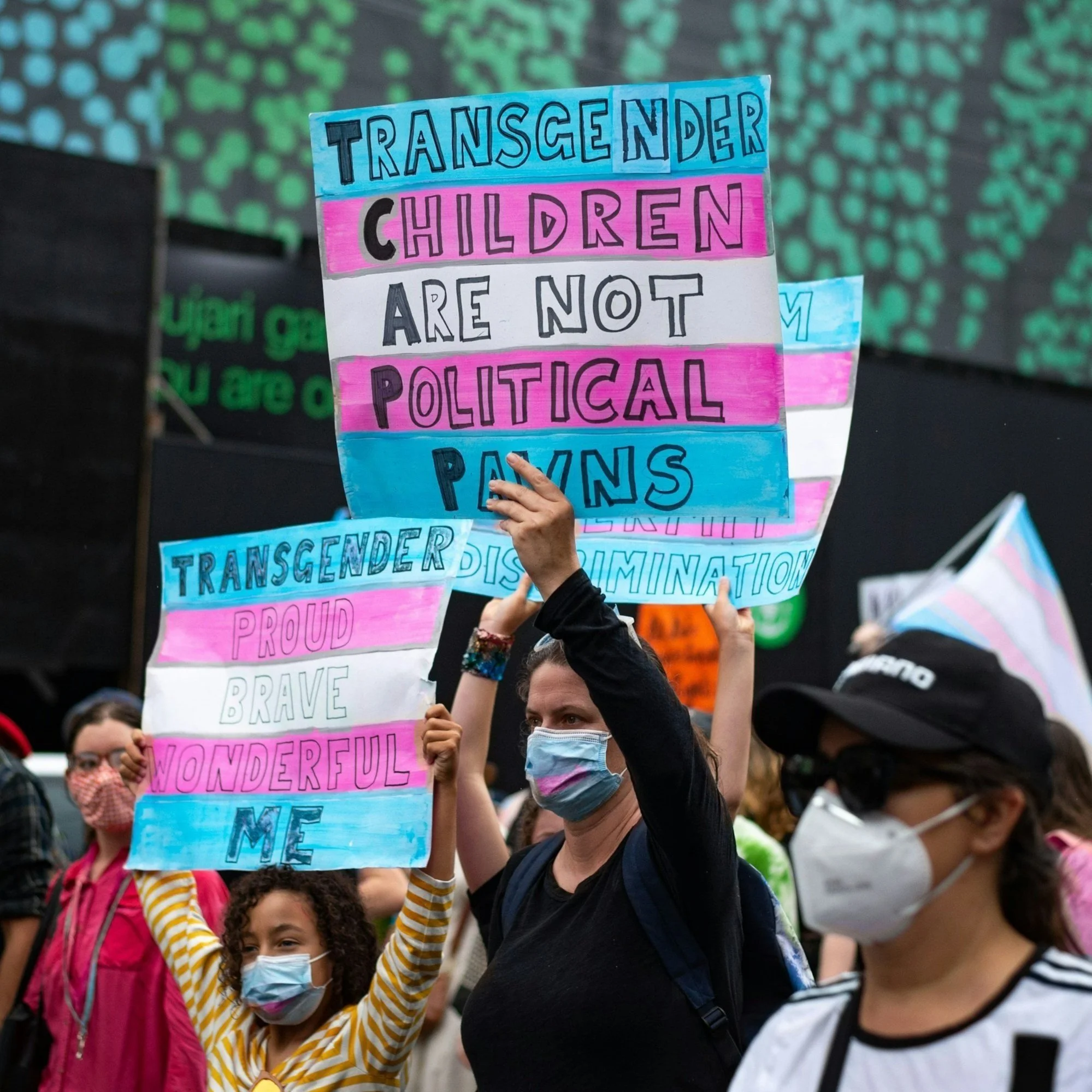 Protestors advocating for transgender children, with signs 'Transgender children are not political pawns,' and 'Transgender proud brave wonderful me.' illustrating Karli K Coaching's values of social advocacy in sex and relationship coaching.
