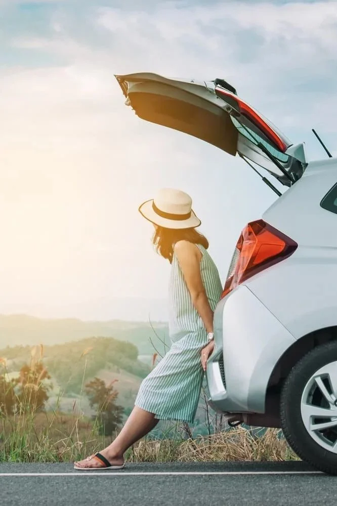 A woman in a striped dress and wide-brimmed hat leaning against the open trunk of a white hatchback car on the side of a rural road, with a scenic landscape of hills and trees in the background during sunset.