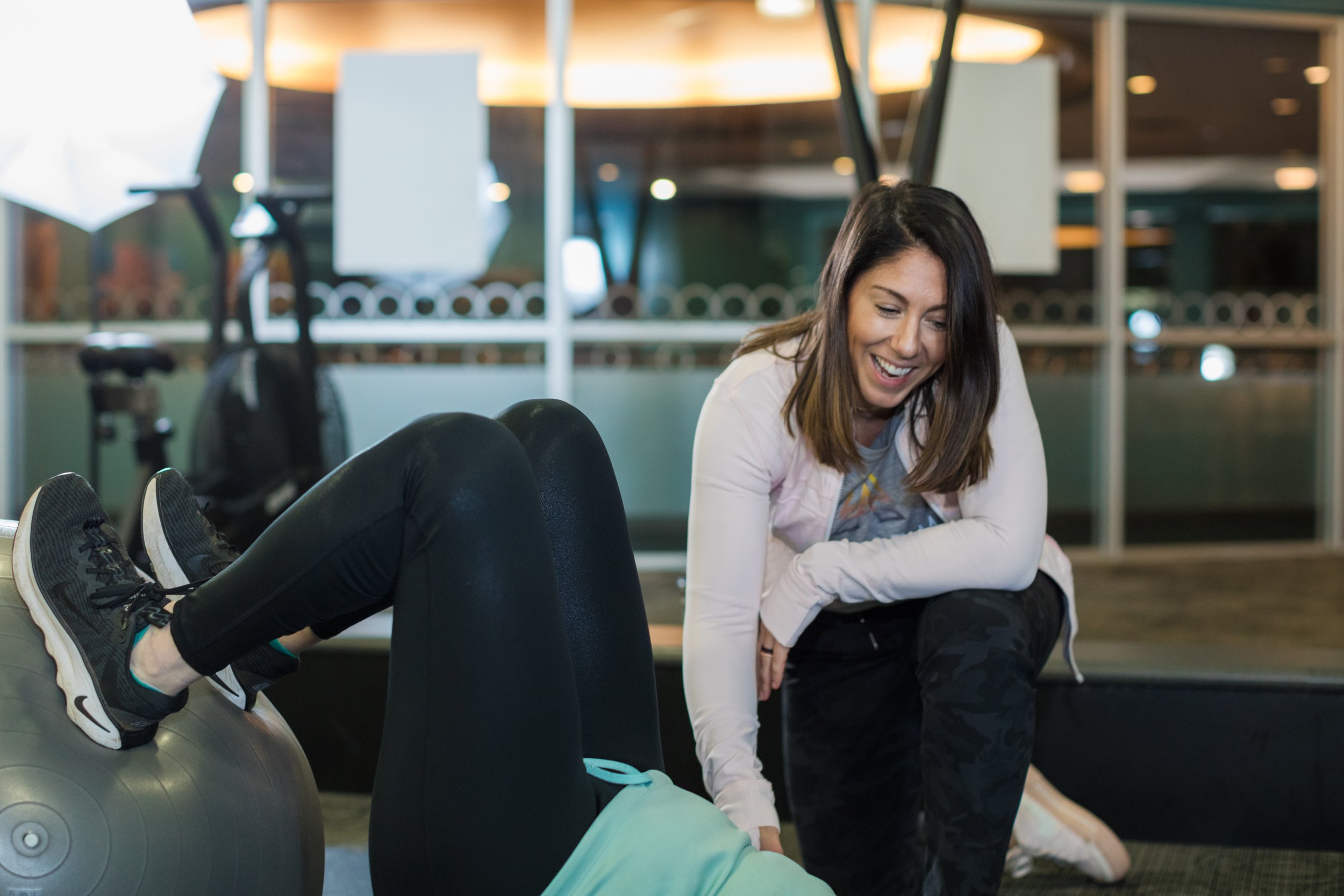A woman with dark hair smiling and assisting another person who is lying on an exercise ball in a gym. The scene is set indoors with gym equipment visible in the background.