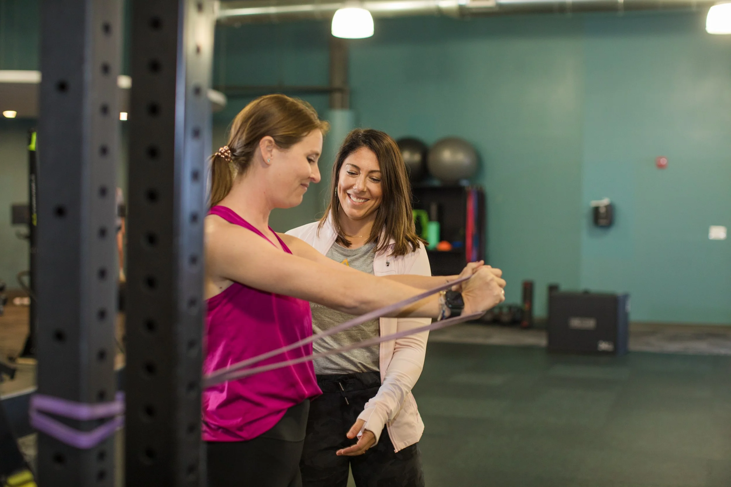 Two women smiling, one in a pink workout top and the other in a light jacket, measuring their arms with a tape measure in a gym.