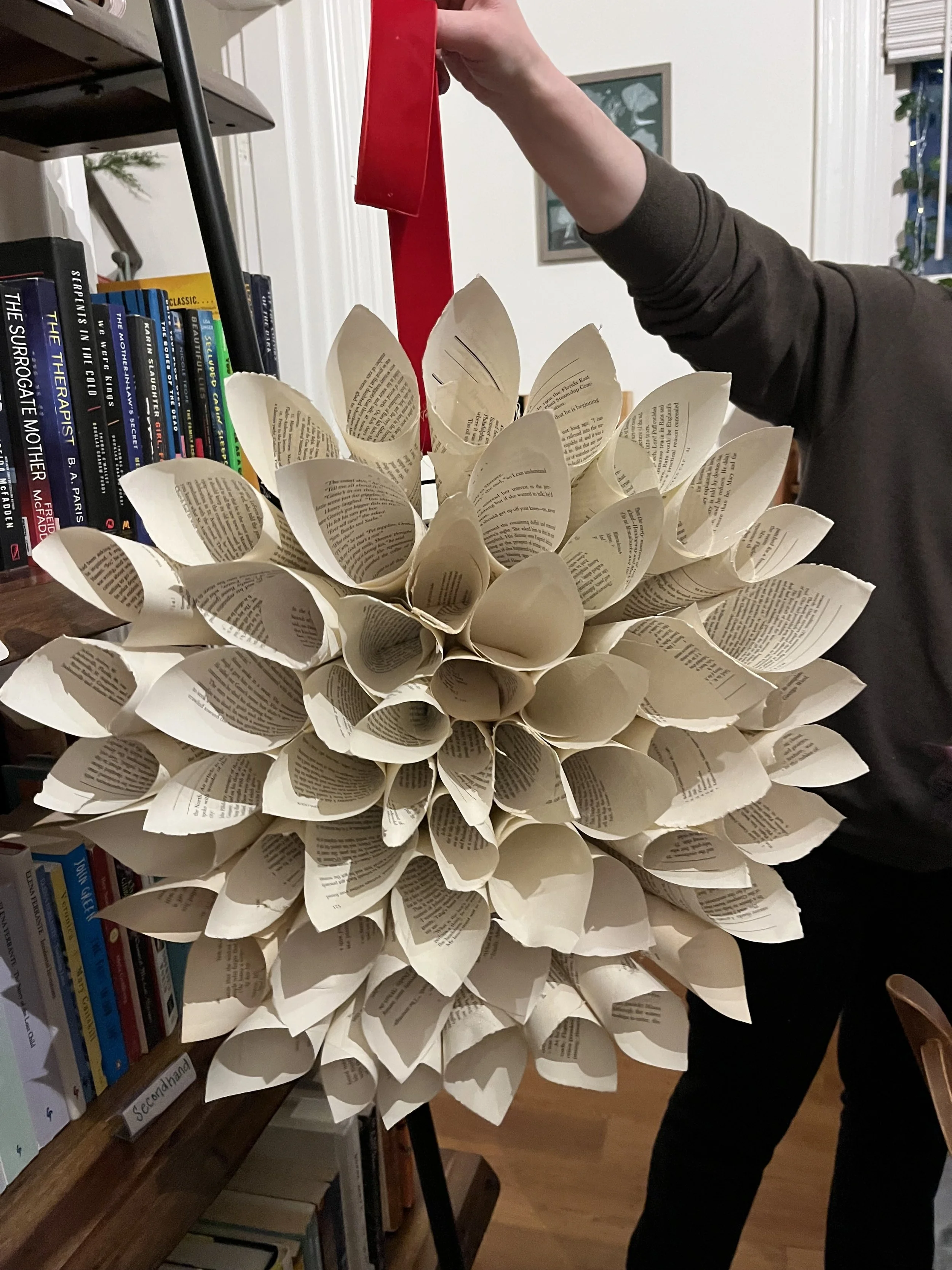 repurposed paper into petals of dahlia wreath transformed into DIY project. person is holding the red ribbon attached to the wreath infront of a book sheld