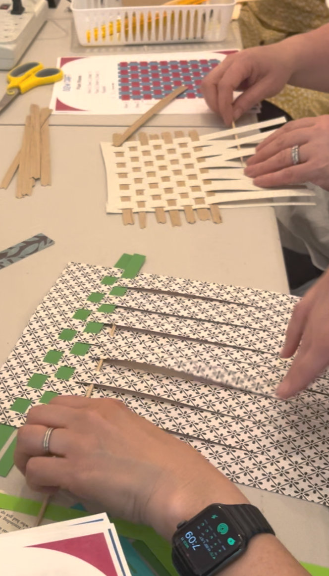 Person weaving paper strips into a square pattern on a table alongside craft supplies and instructions.