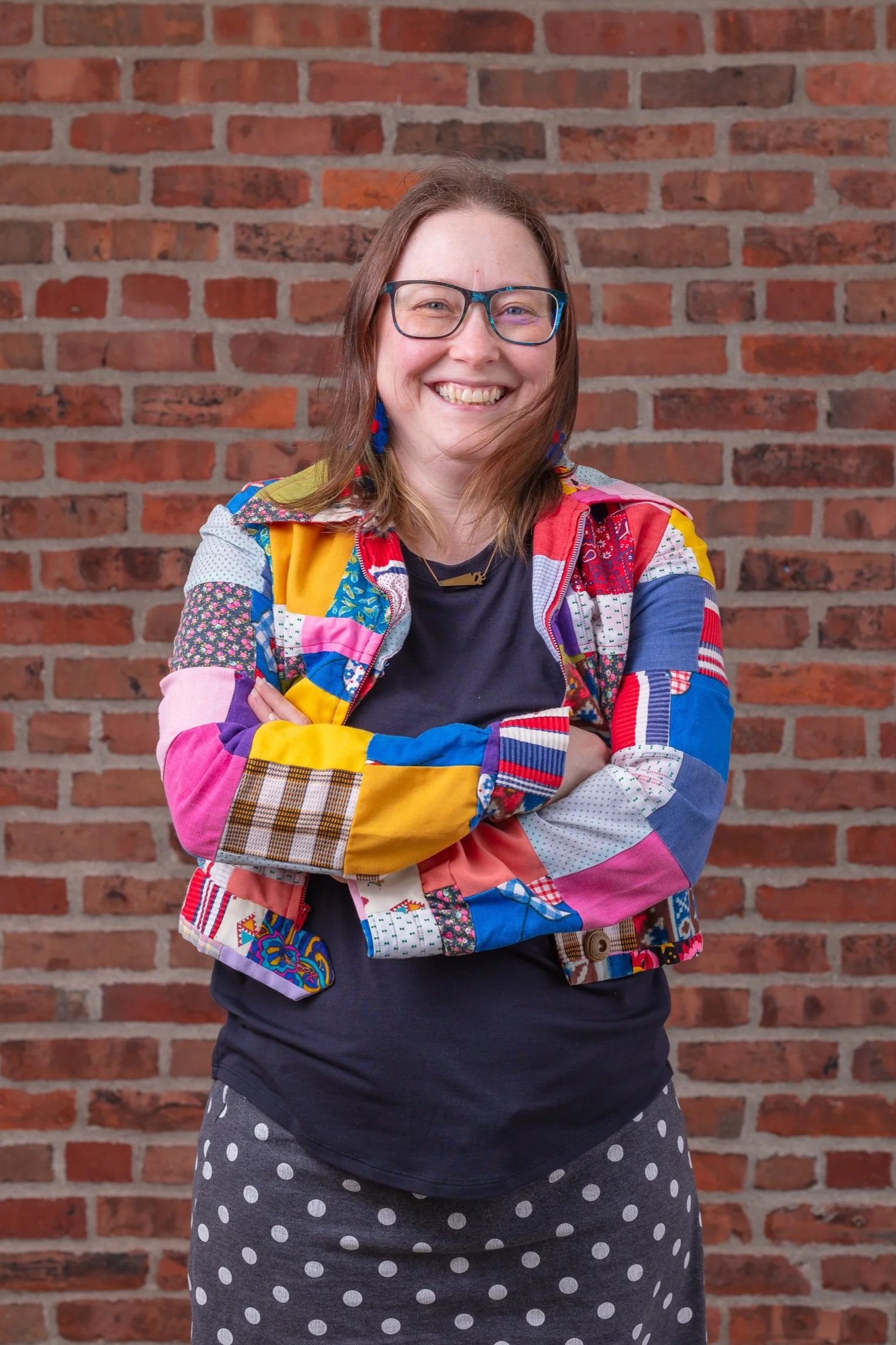 cynthia mcmullen of slow craft llc in patchwork jacket, glasses, smiling in front of red brick wall