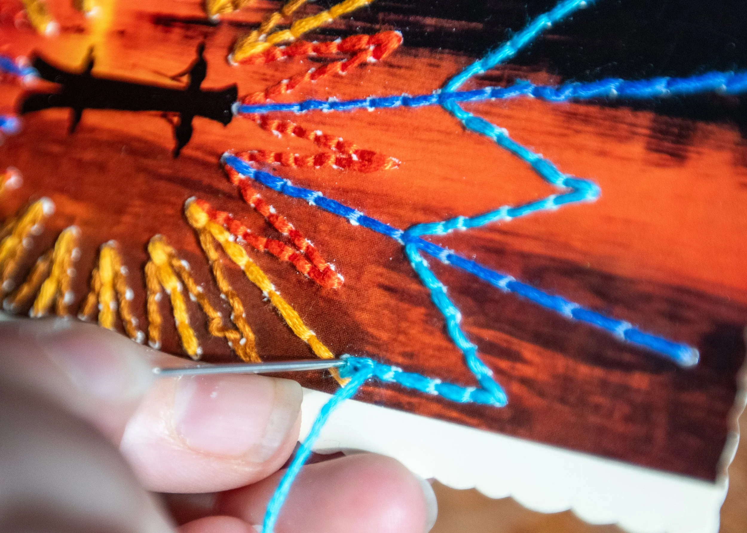 close up fingers poking needle through sunset postcard embroidery