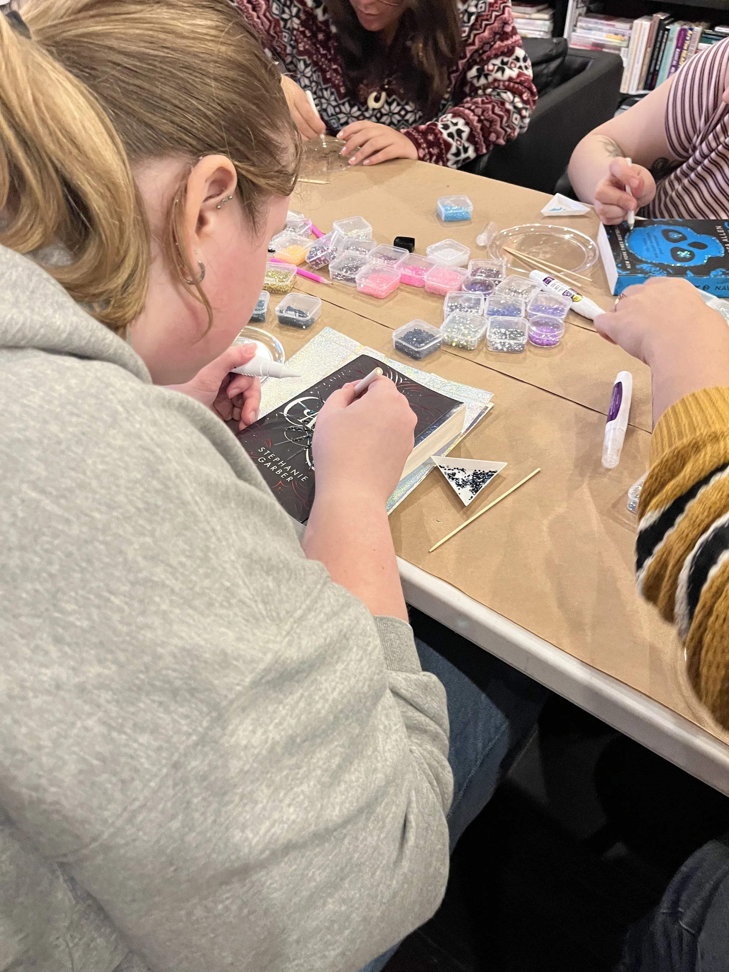 People gathered around a table craft supplies including small containers of beads, a book, and tools, engaged in a bedazzled books activity.