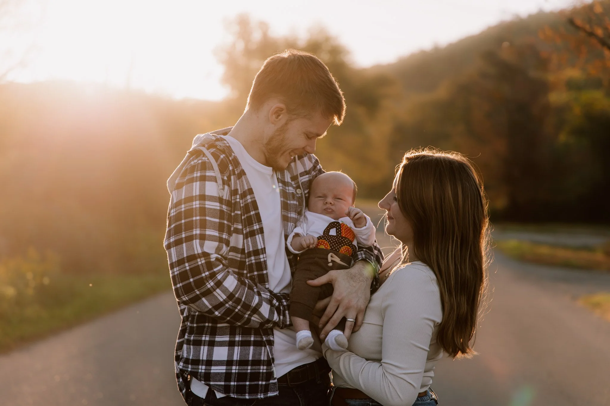 A happy family of three, a man, woman, and baby, outdoors during sunset, with trees and a path in the background.