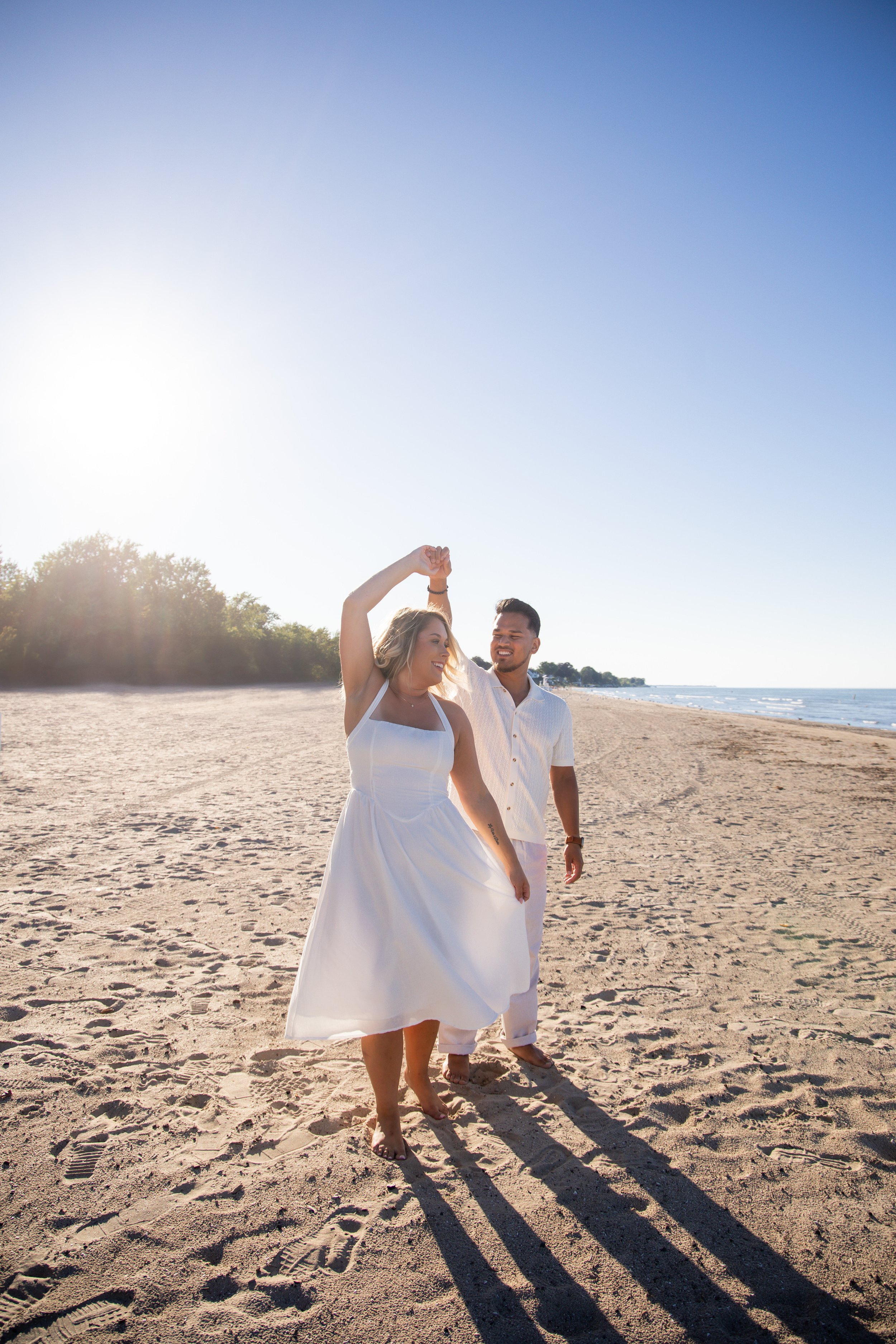 A couple dancing on the beach at sunset, with the woman in a white dress and the man in a white shirt and pants.