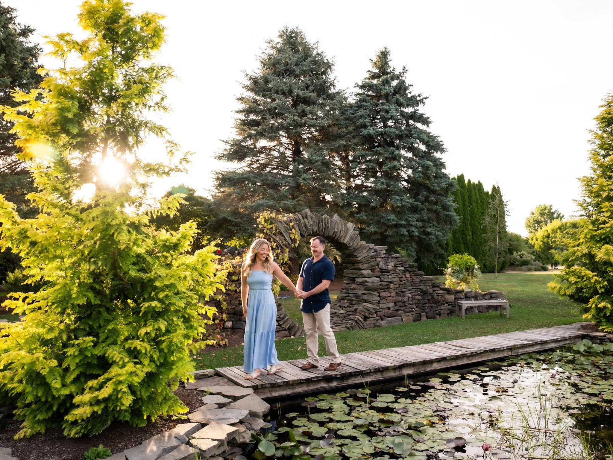 A couple standing on a wooden bridge over a pond with water lilies, holding hands, with trees and a stone arch in the background during sunset.