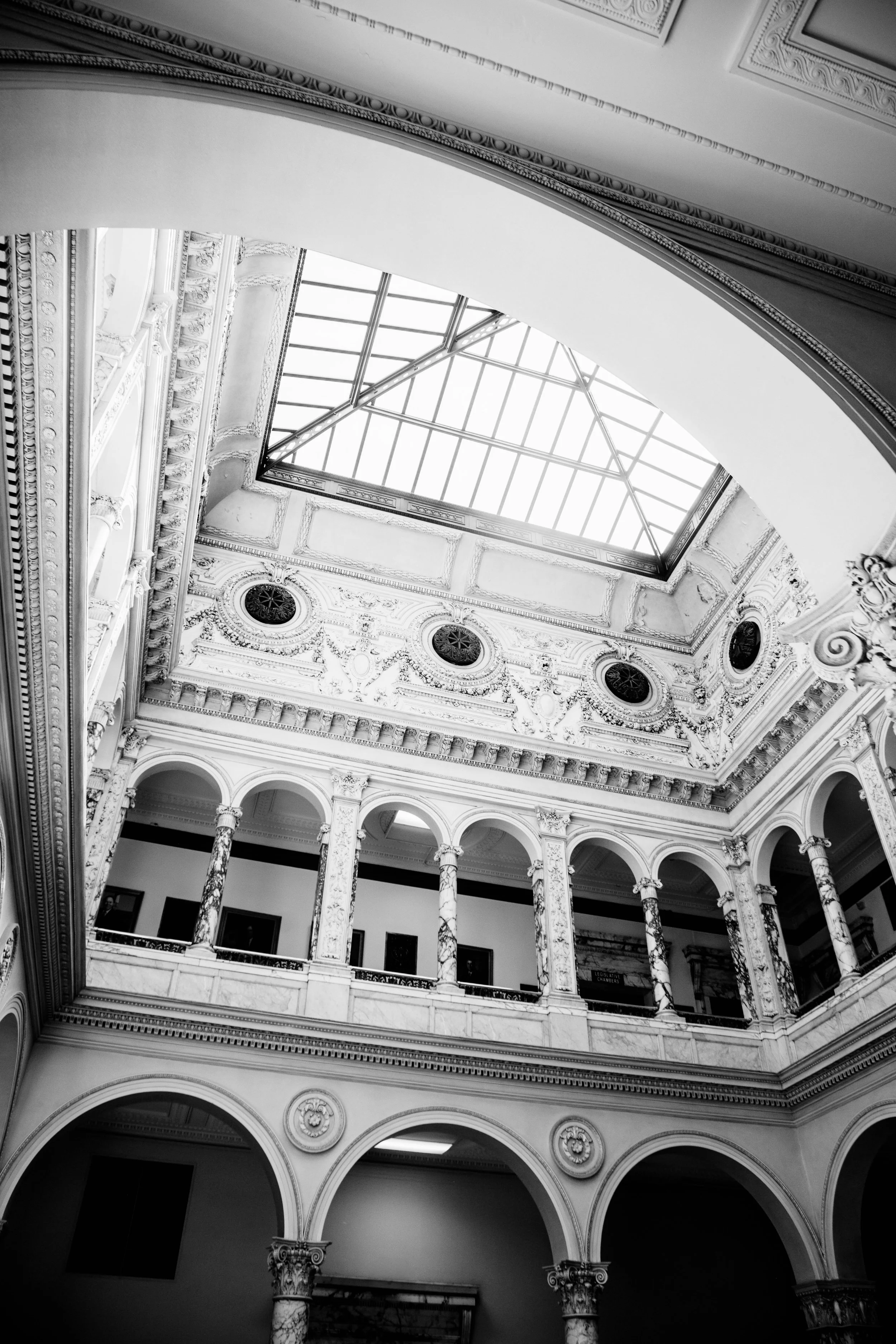 Black and white photo of an ornate interior building with a glass skylight ceiling, arched columns, decorative moldings, and intricate architectural details.