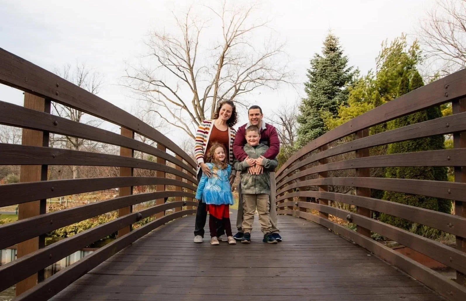 A family of five standing on a wooden bridge outdoors in a park on a cloudy day. The family includes a woman, a man, and three children, all smiling at the camera. The background shows leafless trees and some evergreen trees.