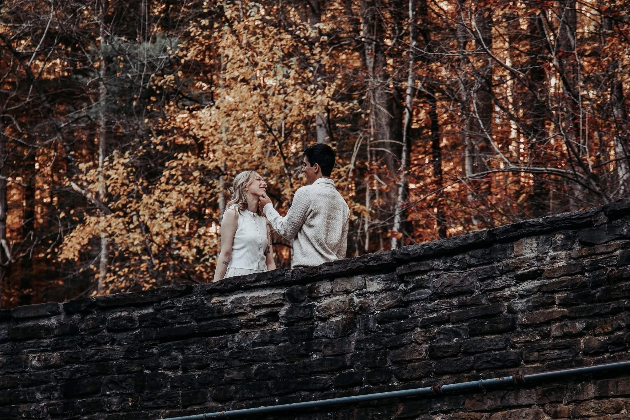 A couple standing on a stone wall in a forest with autumn-colored leaves. The woman is smiling and looking at the man, who is gently touching her face.