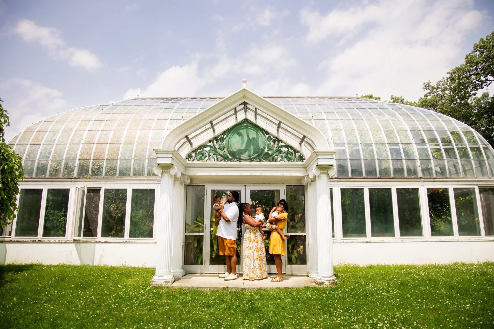 A family of five standing in front of a large glass greenhouse, with two adults and three children dressed in summer clothing.