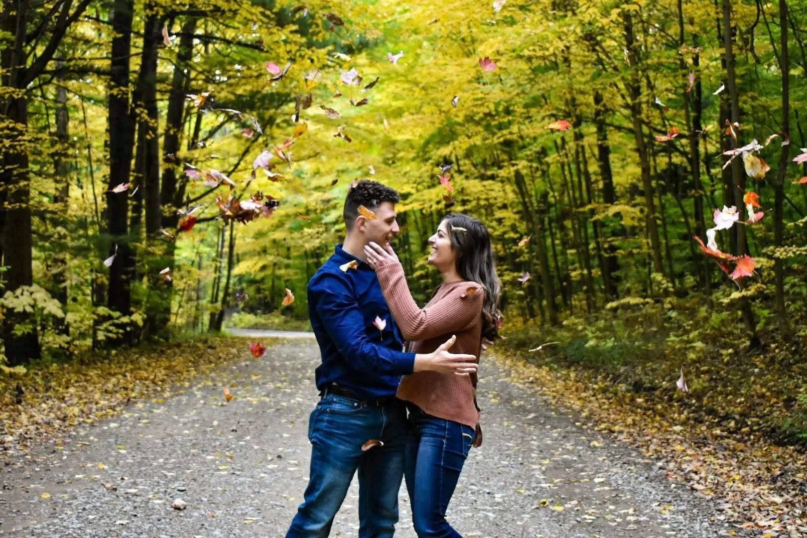 A couple is standing together on a forest path surrounded by yellow autumn leaves and trees, with leaves falling around them as they smile at each other.