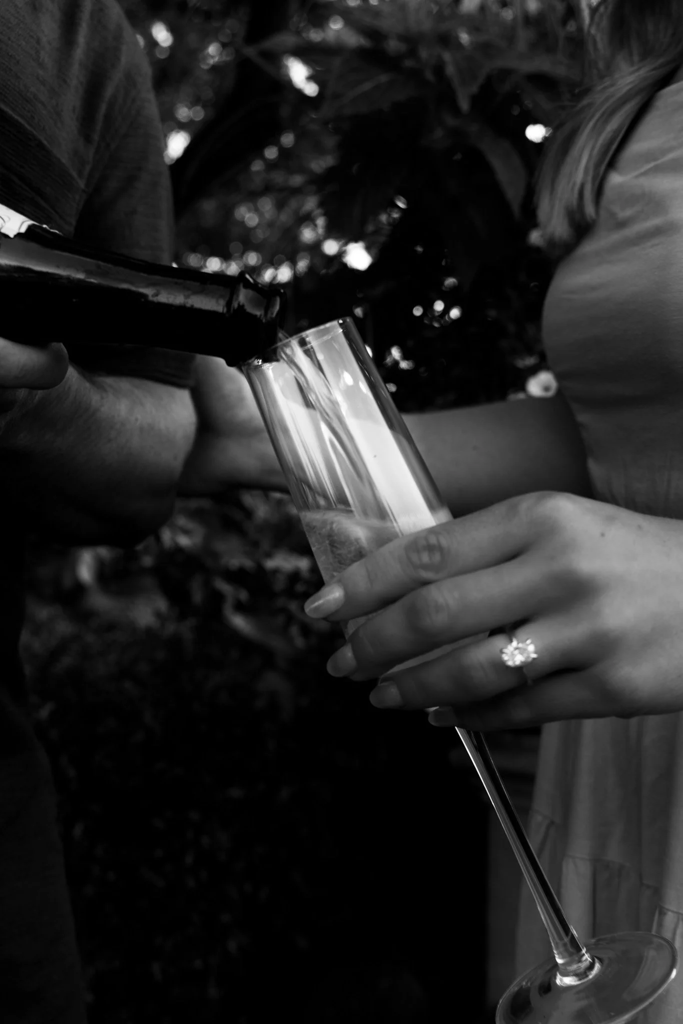 Two people clinking wine glasses outdoors, one pouring a drink into the other's glass in a black-and-white photo.