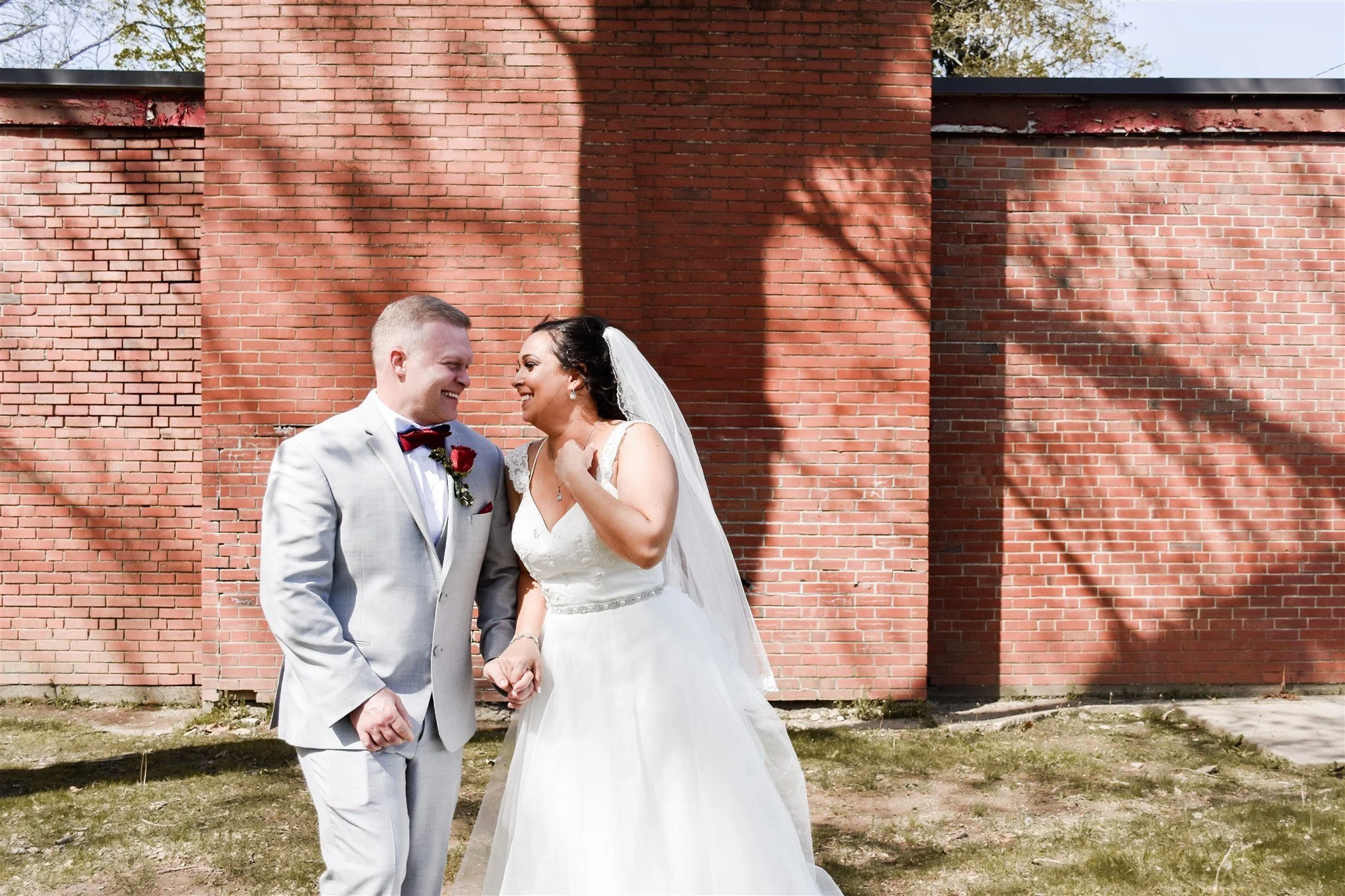 A bride and groom holding hands and smiling at each other outdoors in front of a large brick wall, with shadows cast by nearby trees.