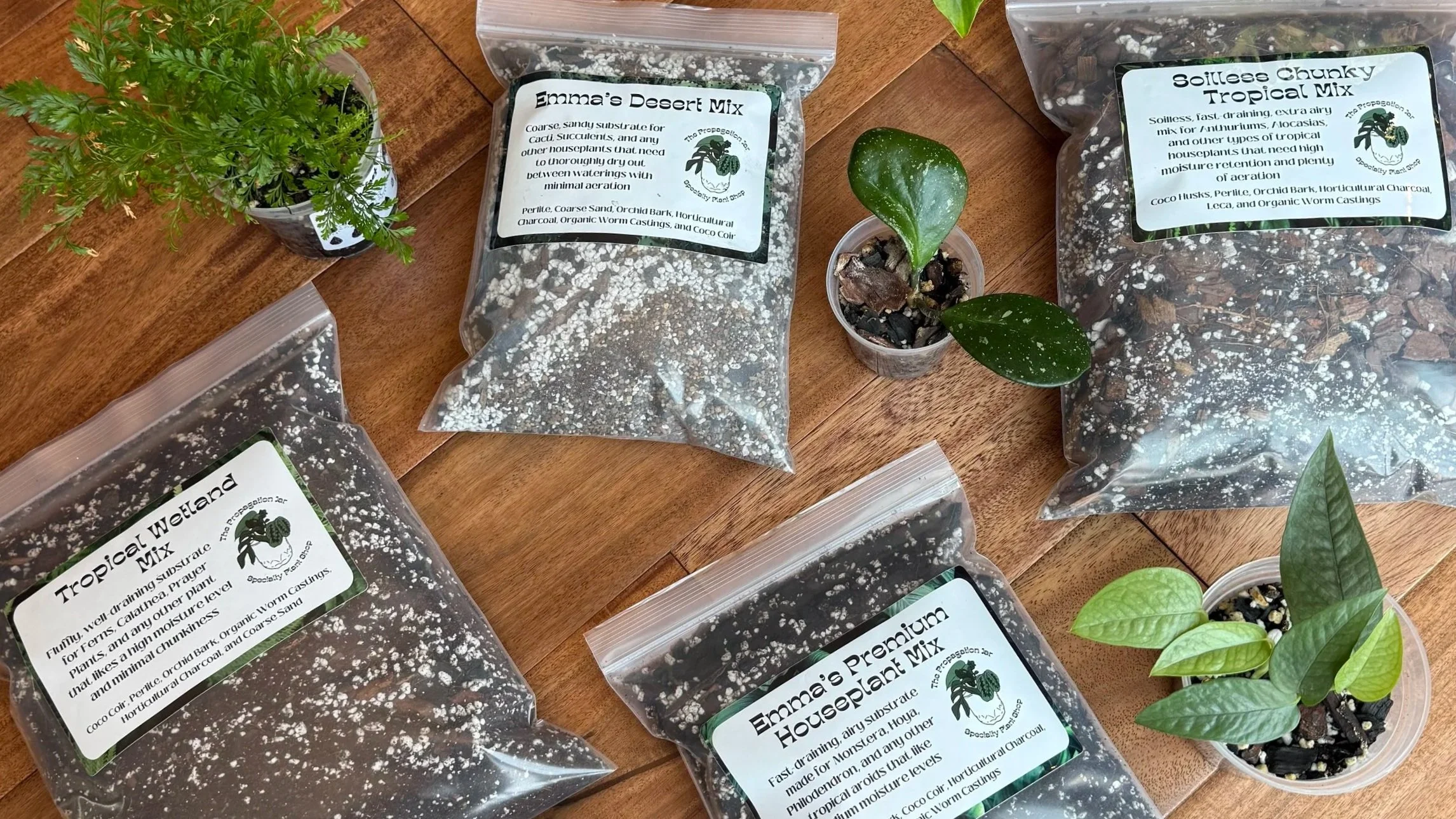 Several clear plastic bags filled with soil and mixed with white particles, labeled with different plant mixes such as "Emma's Desert Mix," "Tropical Wetland Mix," and "Soilless Chunky Tropical Mix," placed on a wooden surface alongside potted plants