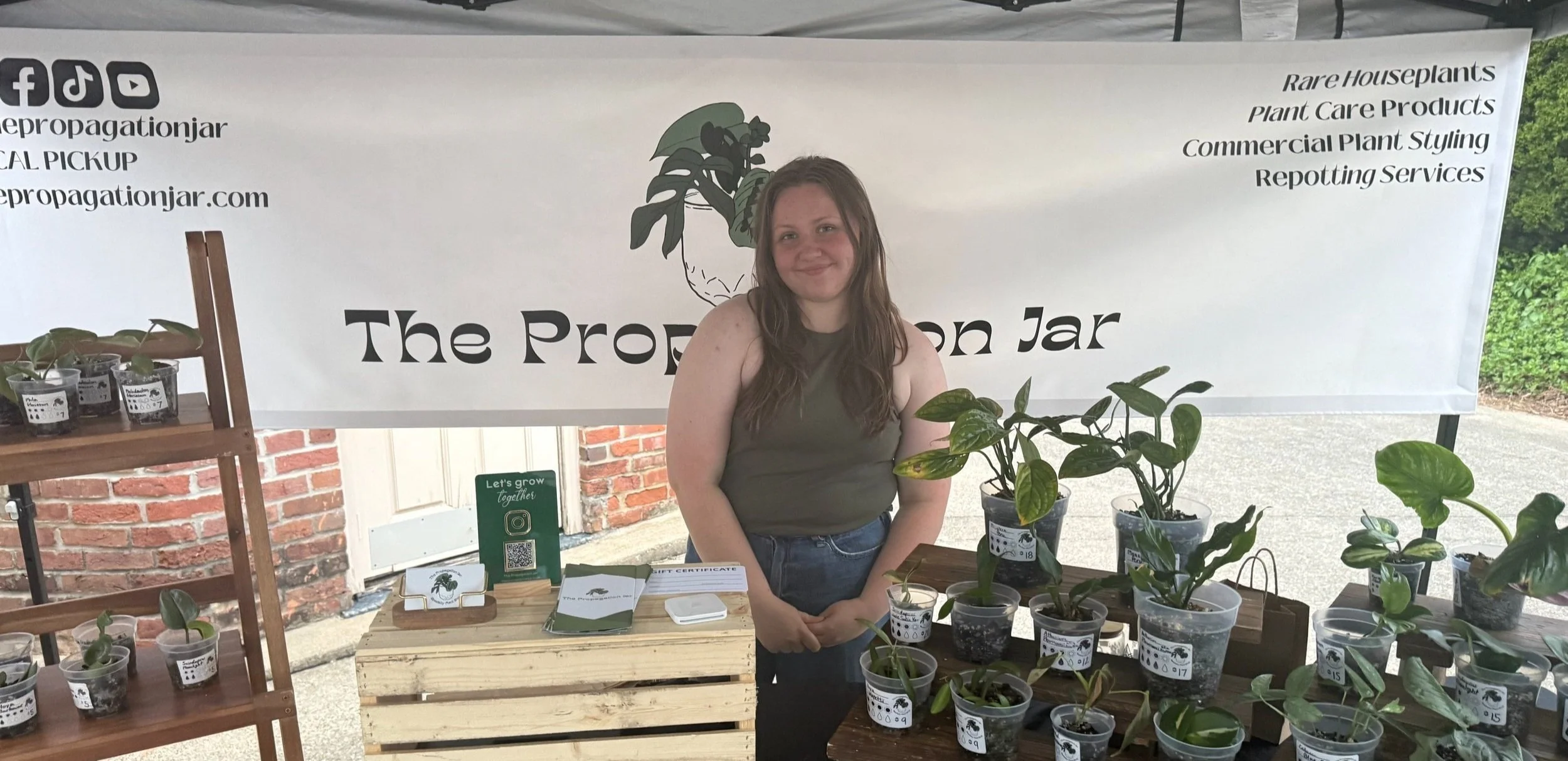 A woman standing at a plant sale booth with potted plants on tables and shelves, and a banner behind her advertising rare houseplants and plant care products.