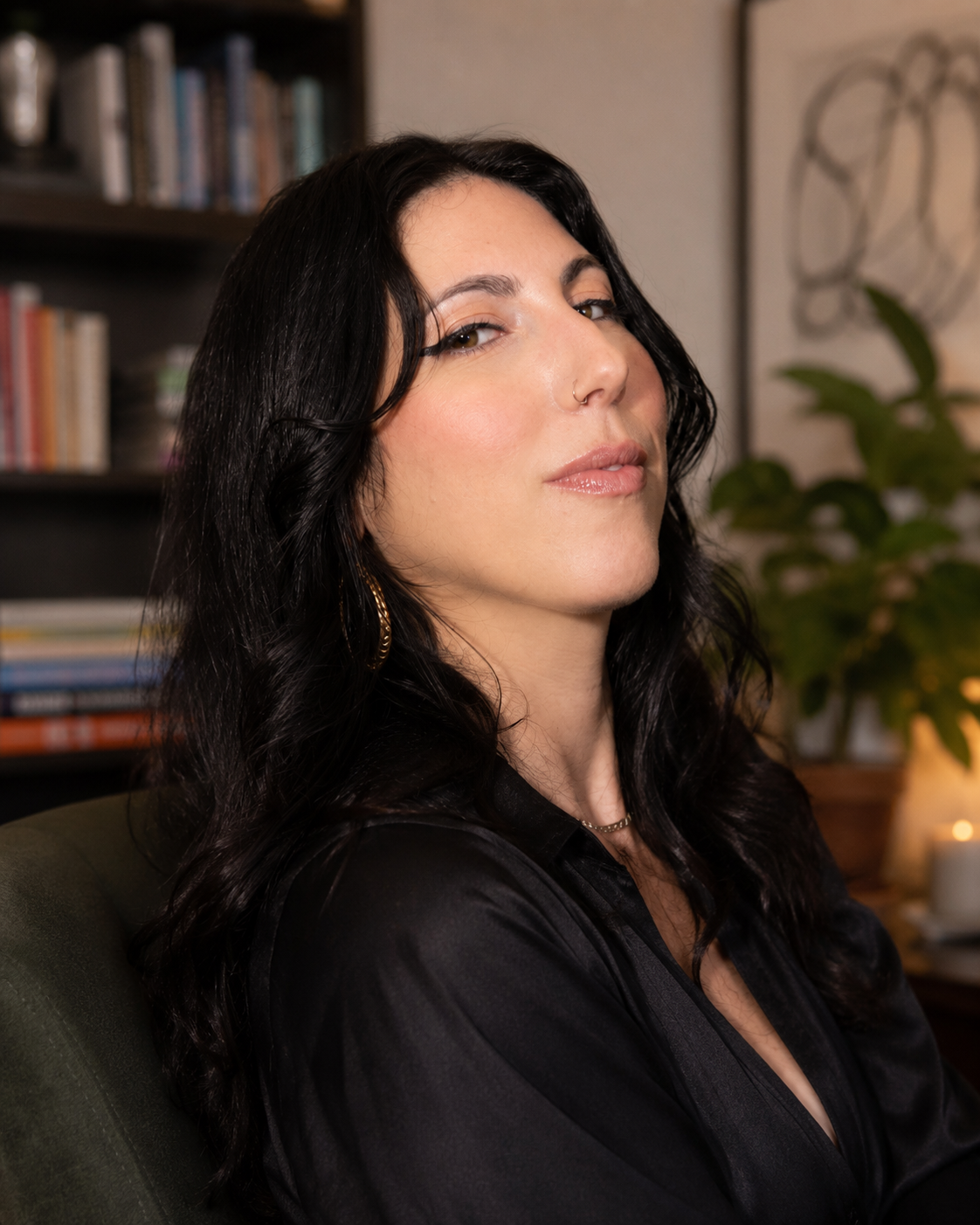 Portrait of Sarabeth Linden, LMHC. Description: a woman with long curly black hair, red lipstick, winged eyeliner, and a nose ring smiling at the camera, sitting indoors with yellow walls and a colorful poster in the background.