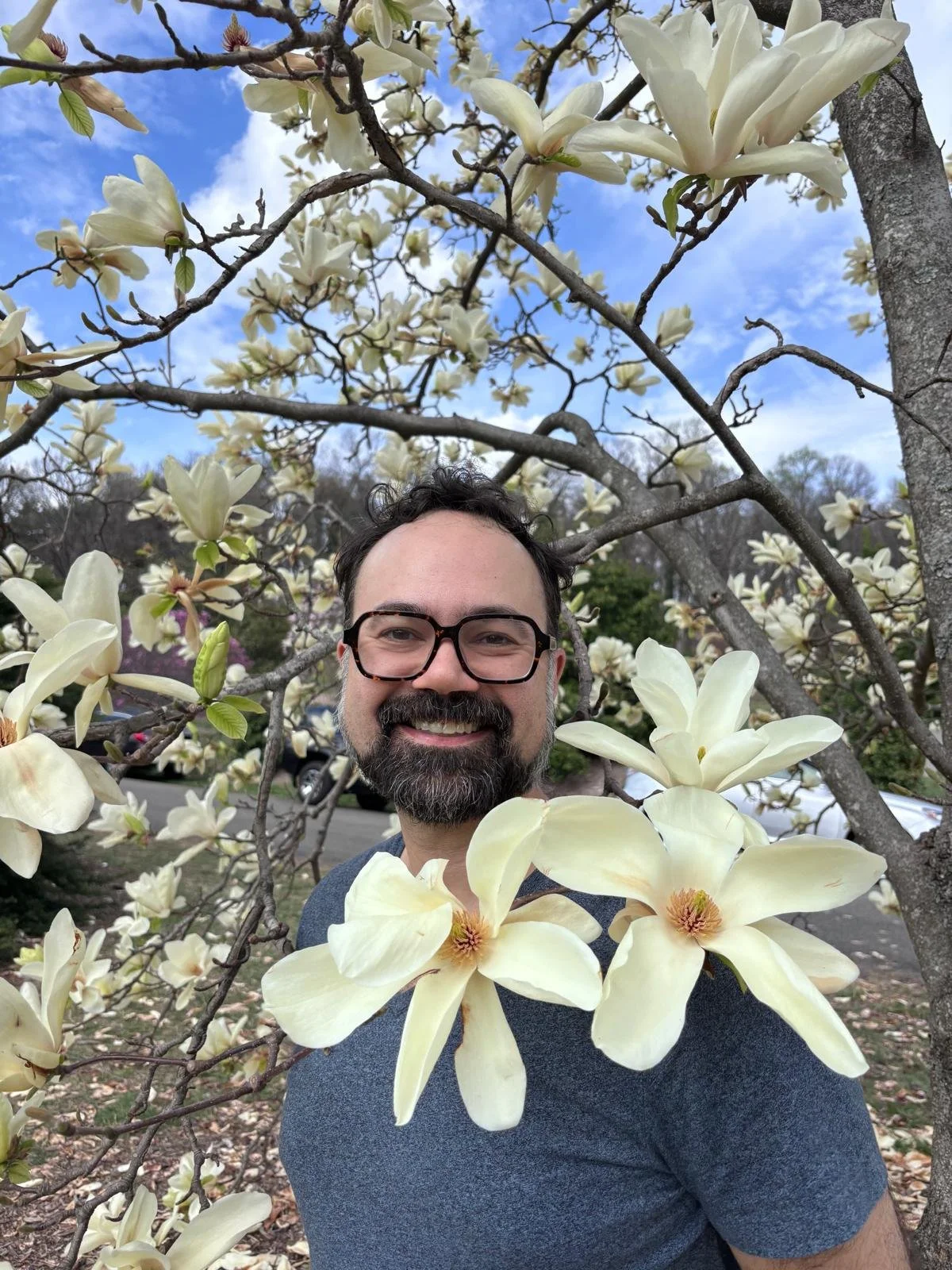 A smiling man with dark hair, glasses, and a beard stand in front of a blooming magnolia tree, with large cream-colored flowers in the foreground.